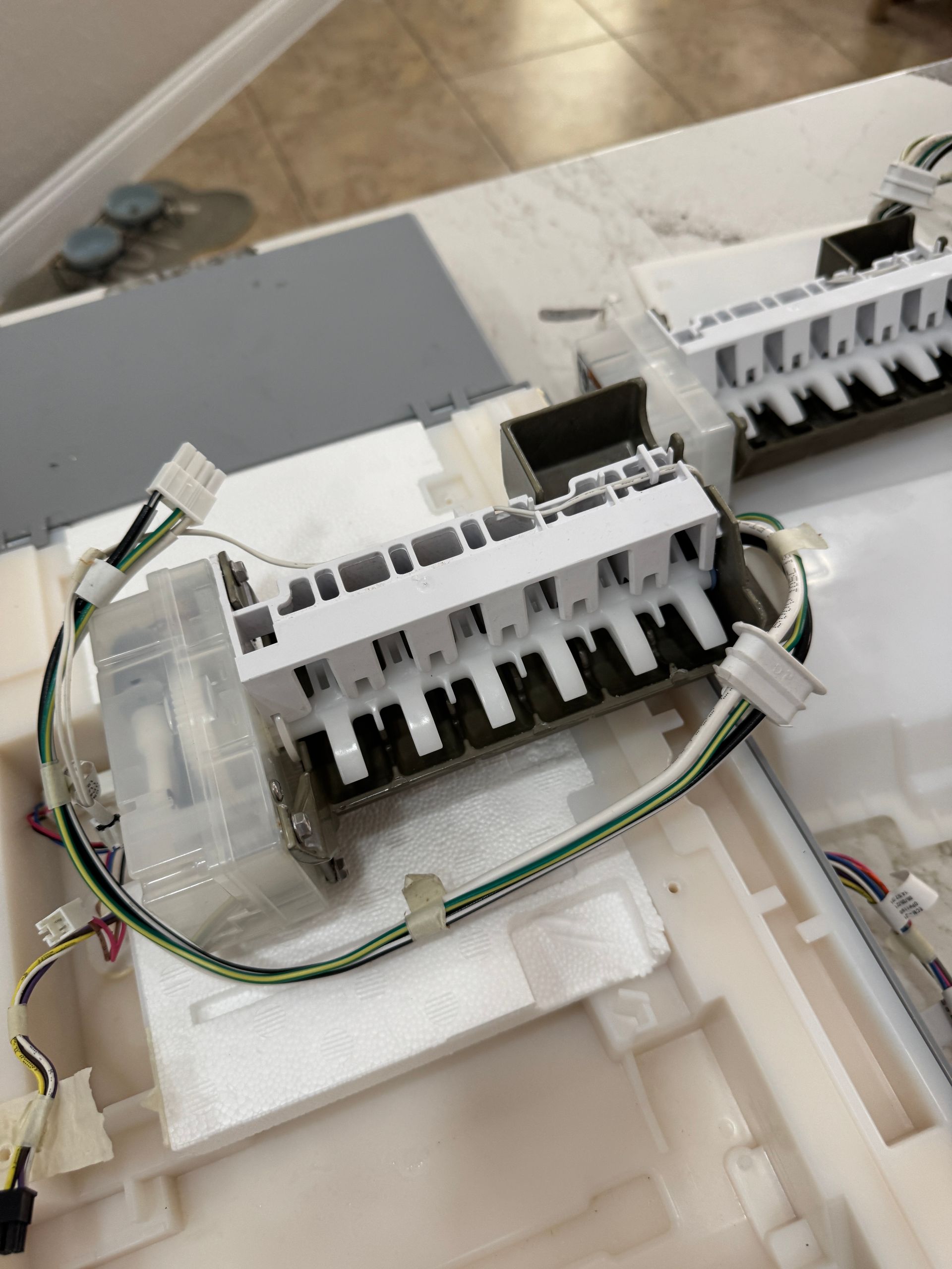 Disassembled white appliance ice maker components with wiring, on a white surface.