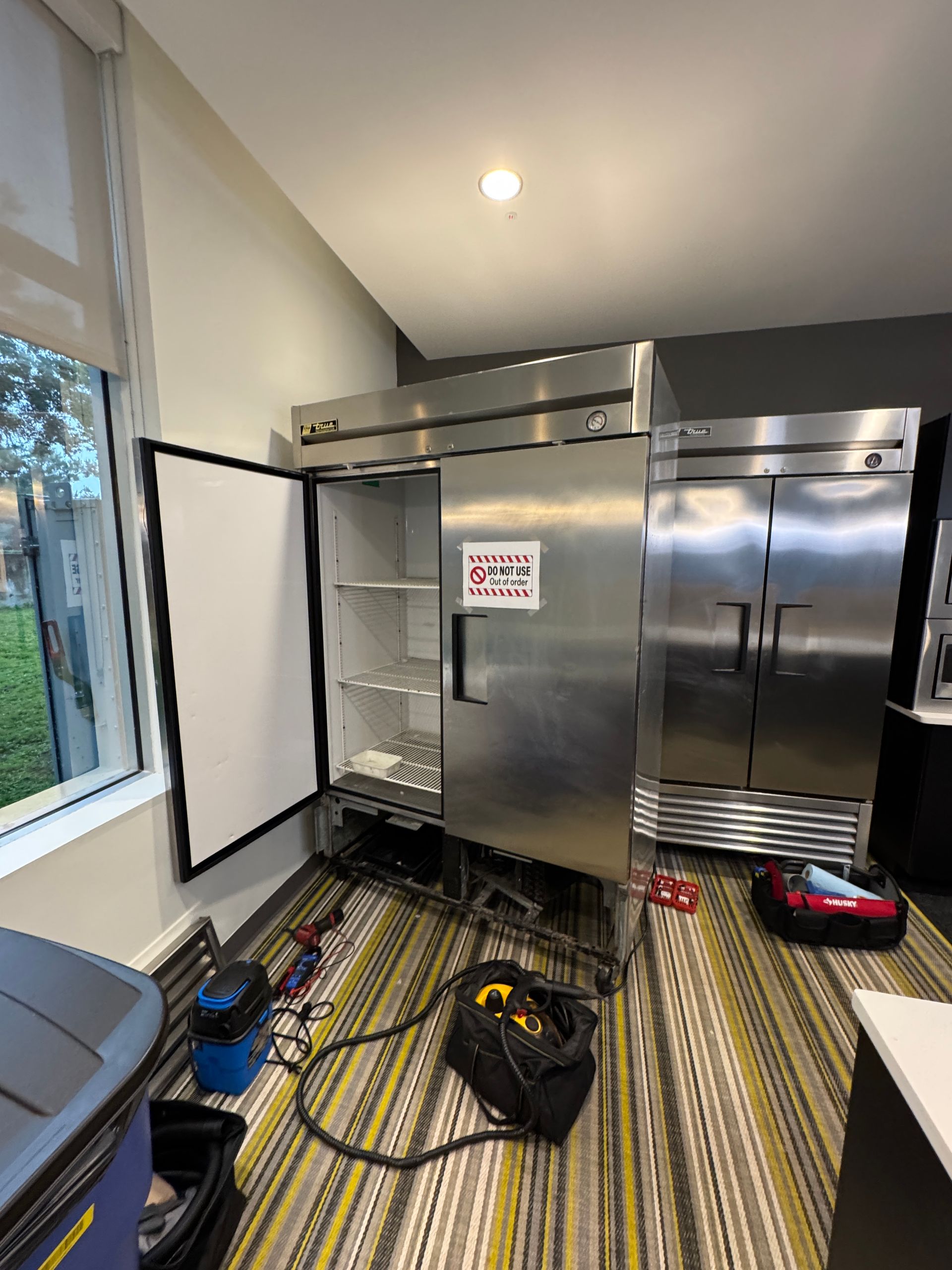 A commercial refrigerator with the door open, tools on the floor. Interior setting, well-lit.