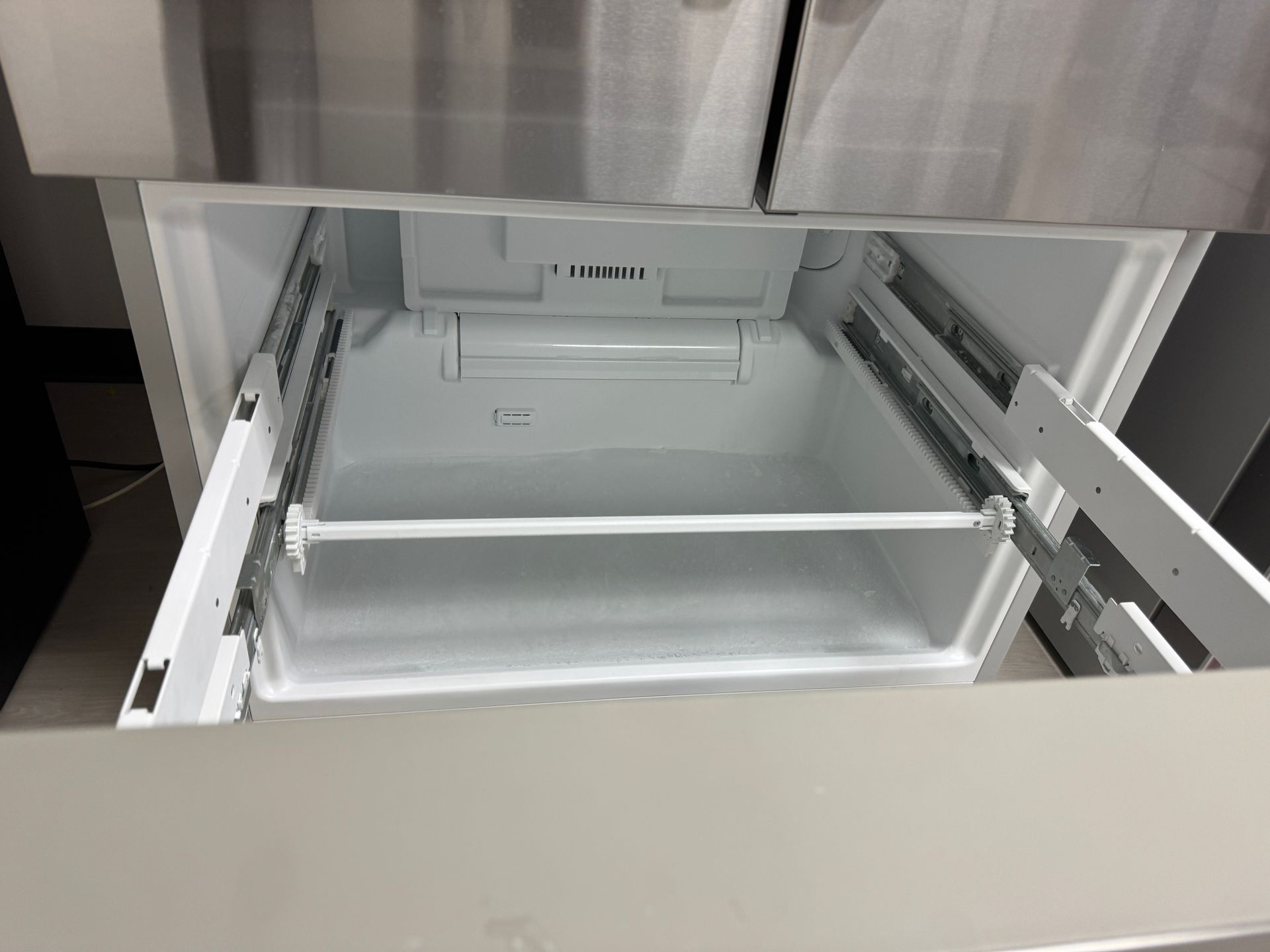 Open refrigerator freezer drawer with white interior and silver rails.