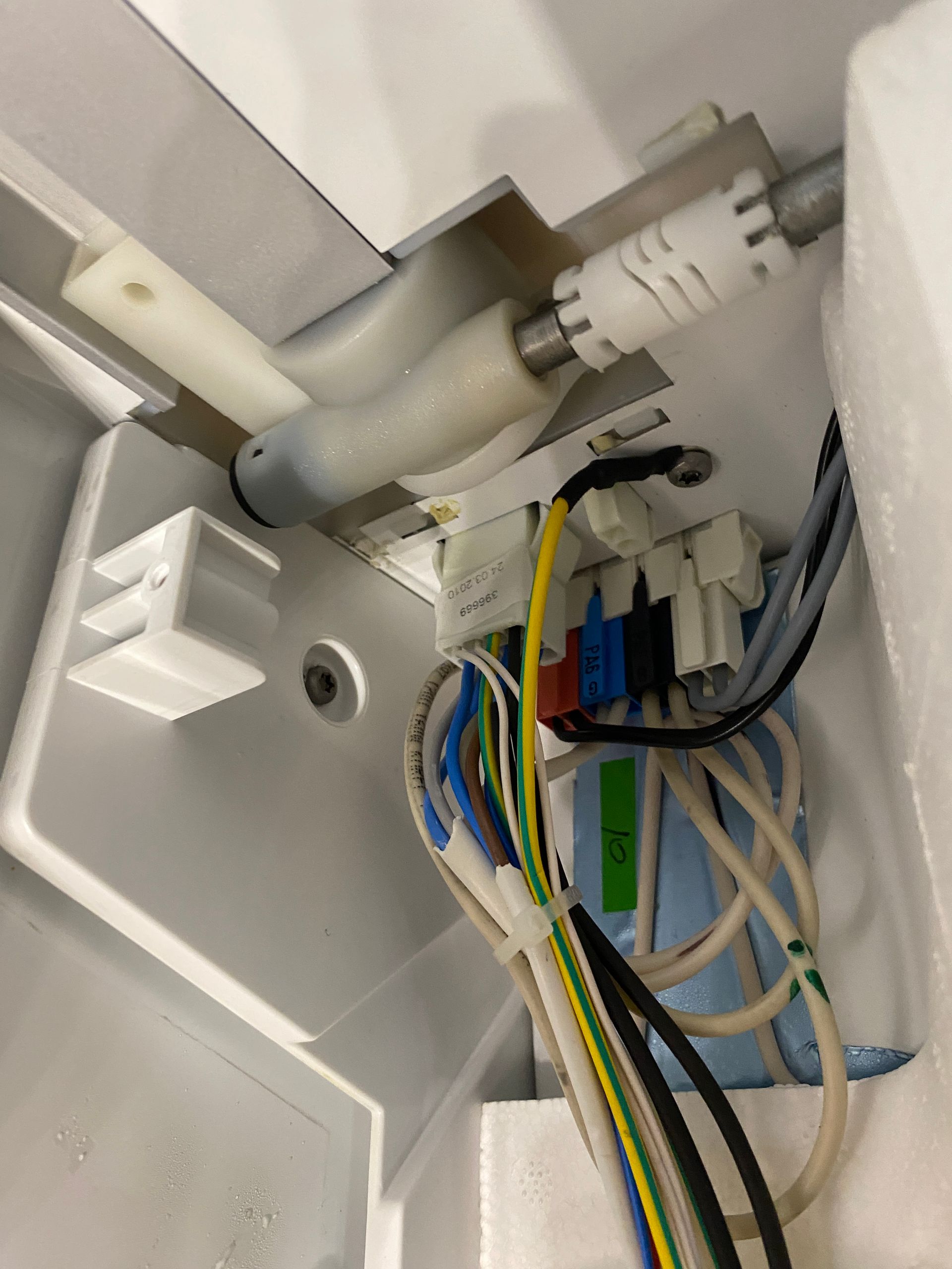 Inside of a refrigerator: Ice maker mechanism with white plastic parts and a bundle of colored wires.
