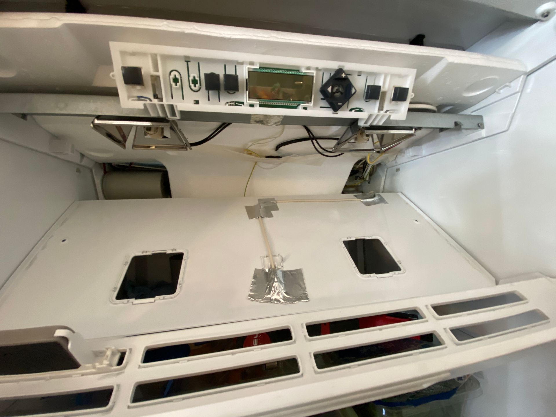 Interior of a refrigerator, with upper panel removed, exposing wiring and electronics. White walls and ventilation slits.