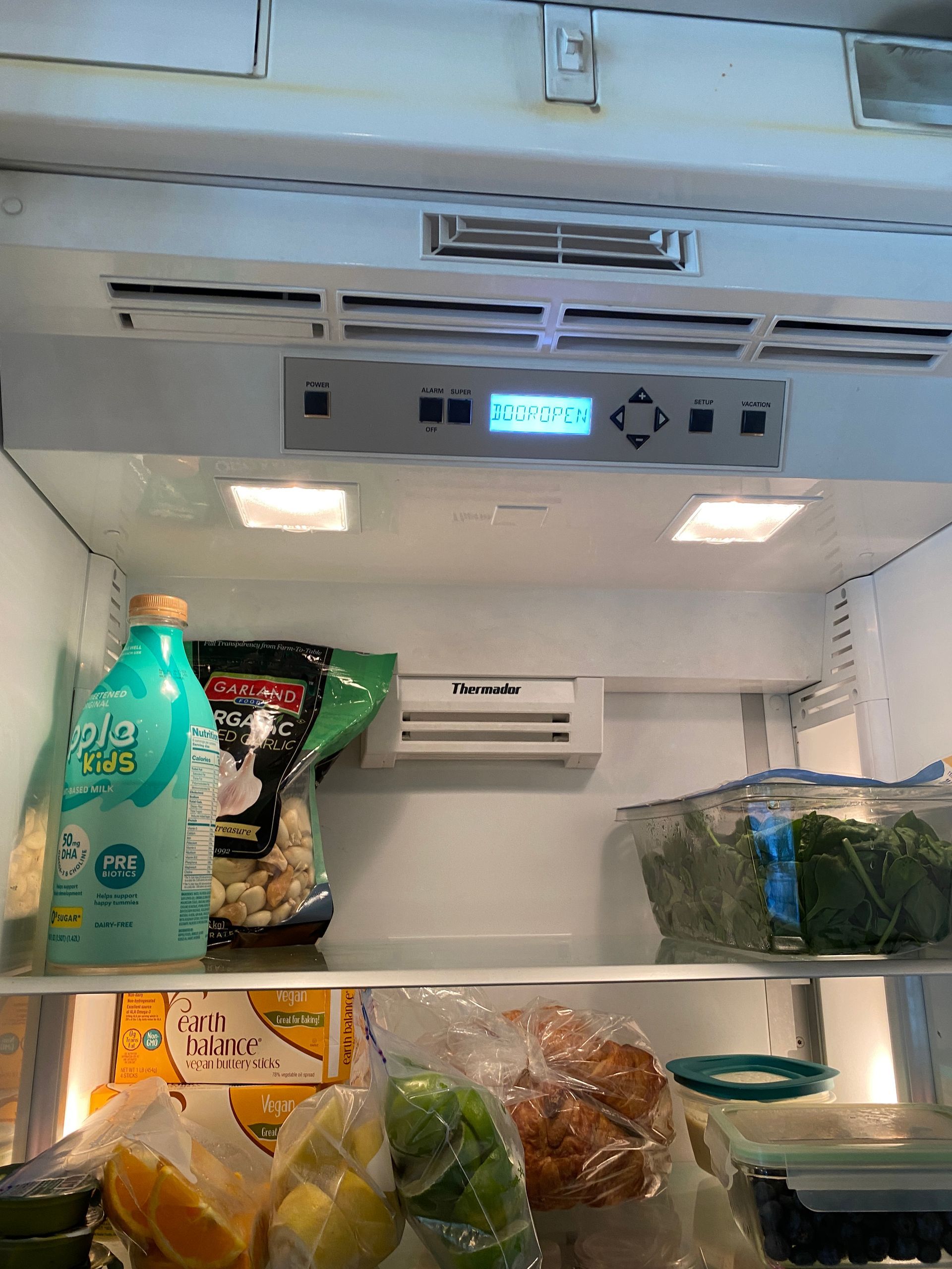 Open refrigerator with food, including milk, vegetables, and fruit. Digital control panel visible.