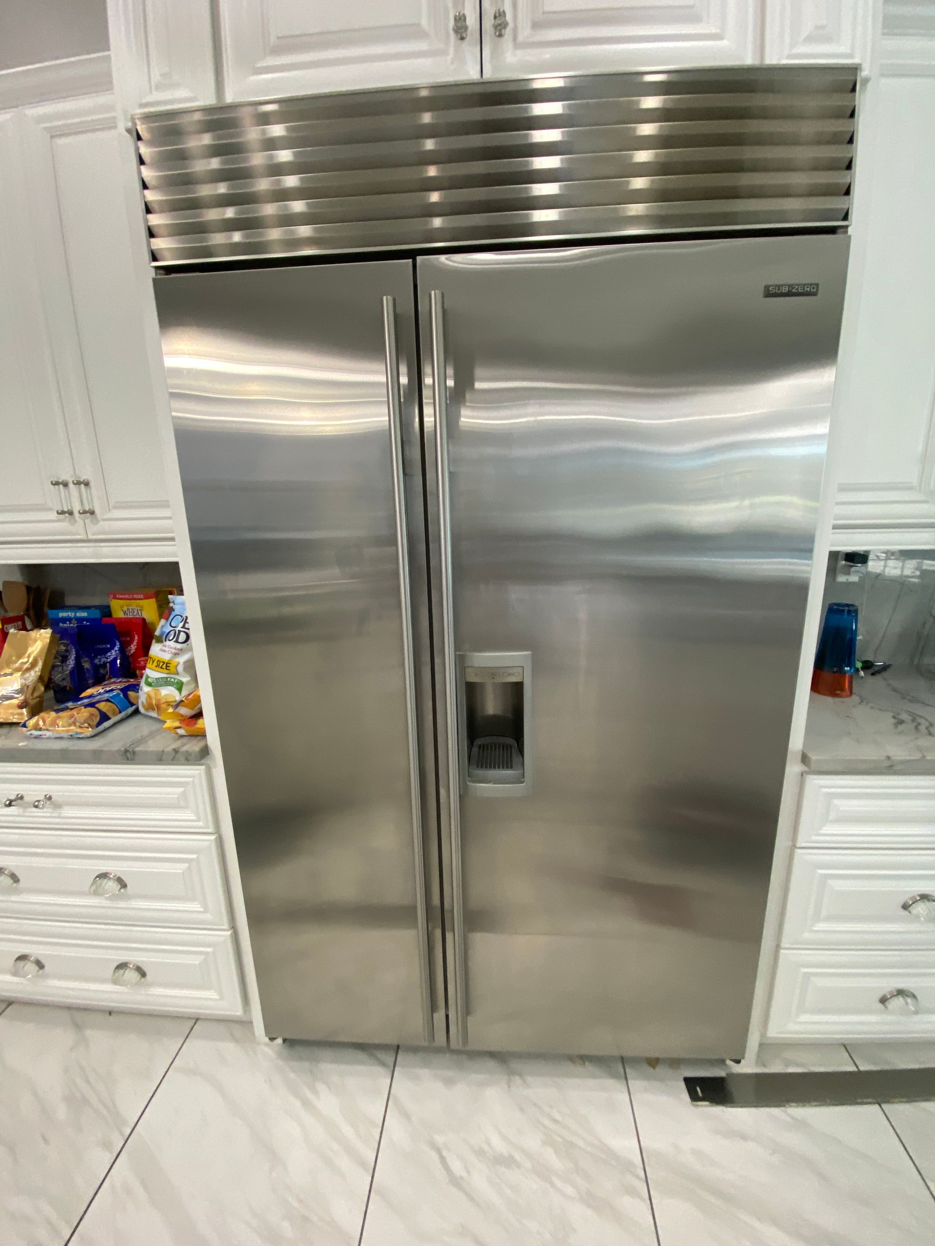 Stainless steel side-by-side refrigerator in a white kitchen, flanked by white cabinets and drawers.