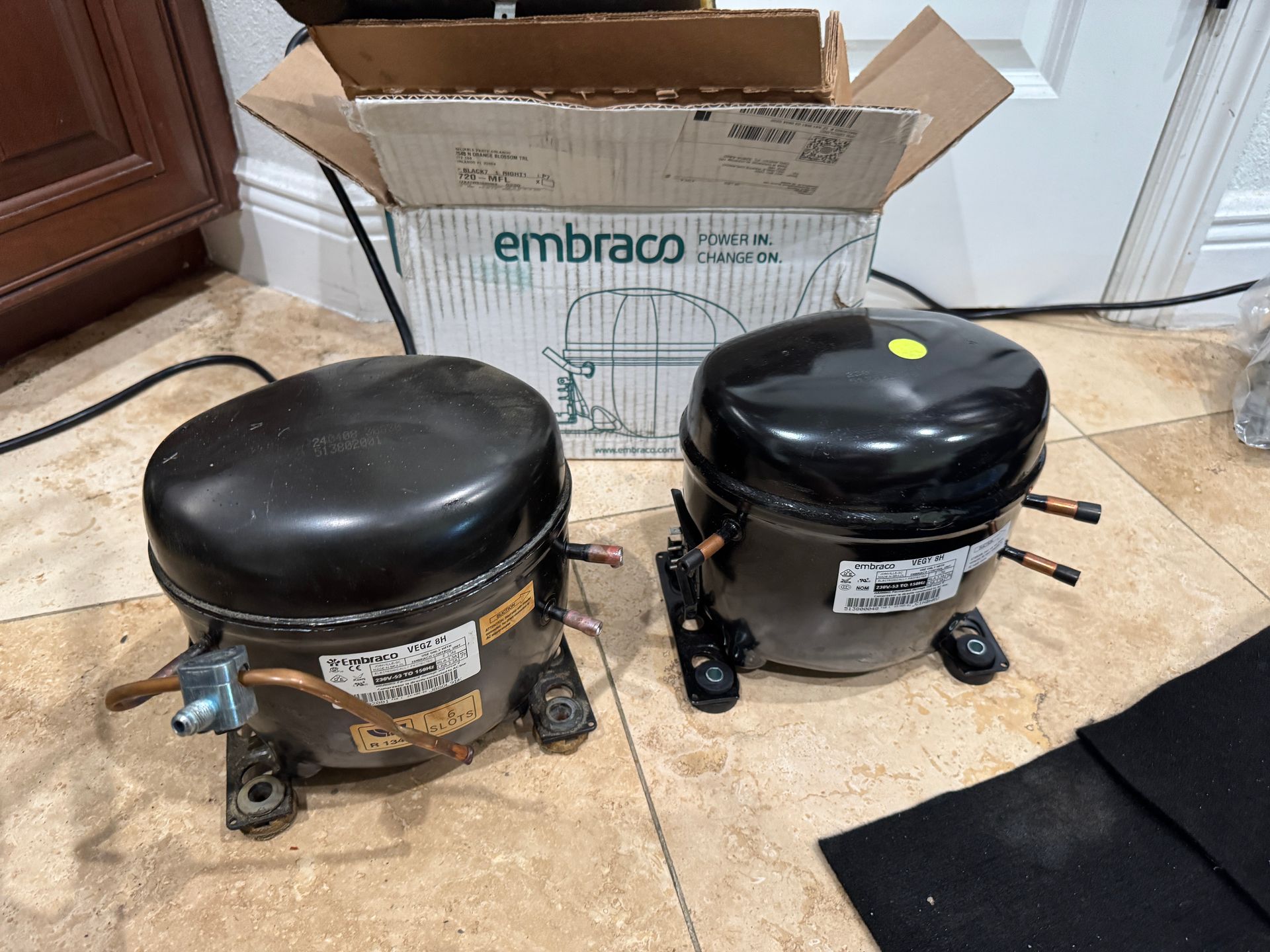 Two black Embraco refrigerator compressors next to a cardboard box on a floor.