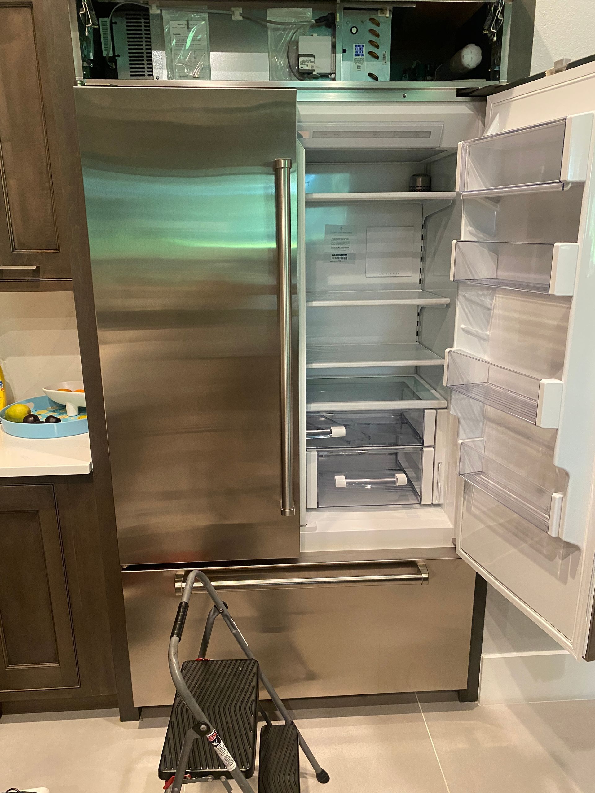 Stainless steel refrigerator with open doors, step stool in front. Kitchen setting.
