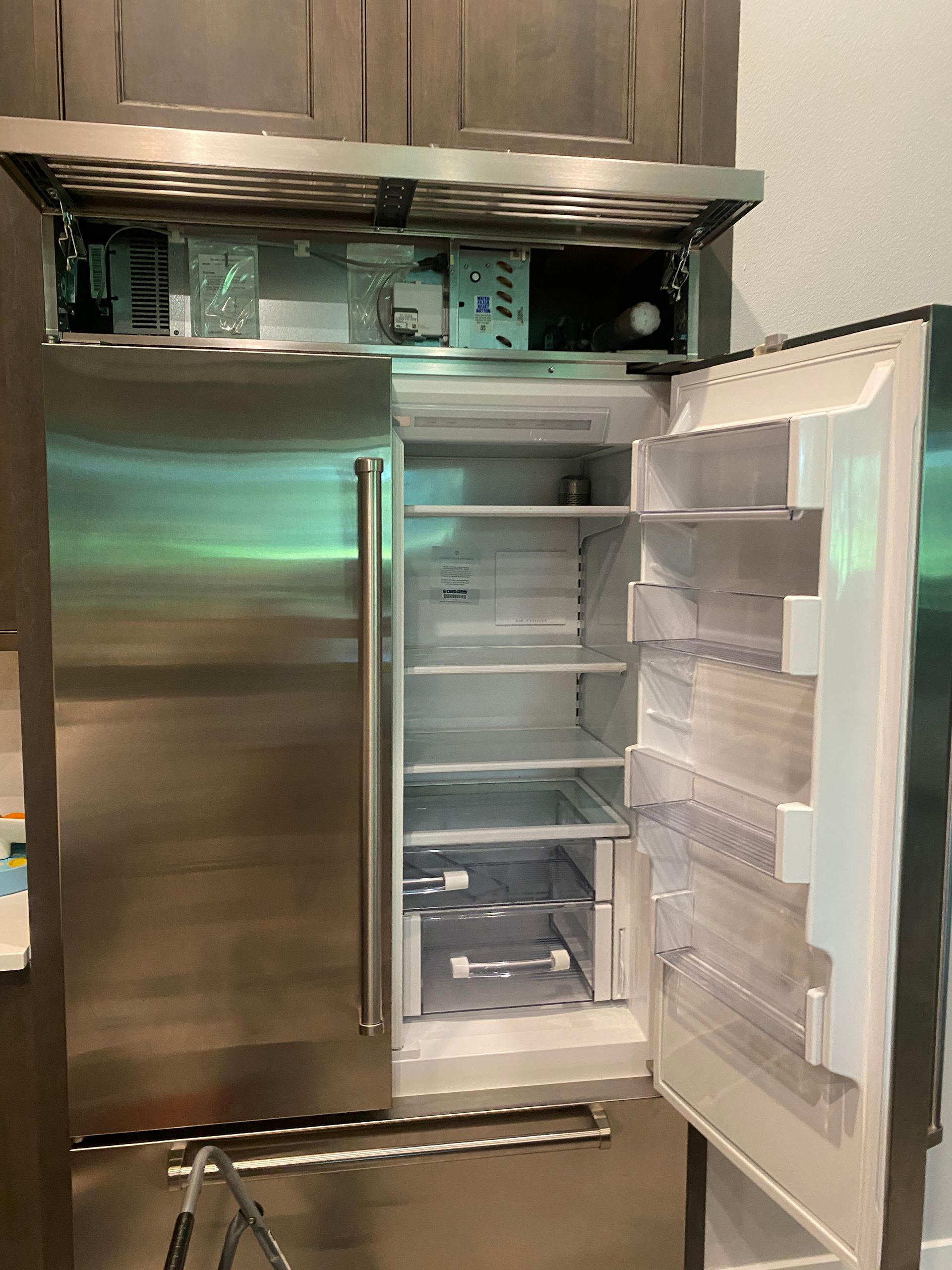 Stainless steel refrigerator, open, revealing empty shelves. Interior components visible above.