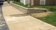 Concrete sidewalk beside a brick house with a low retaining wall and grass strip
