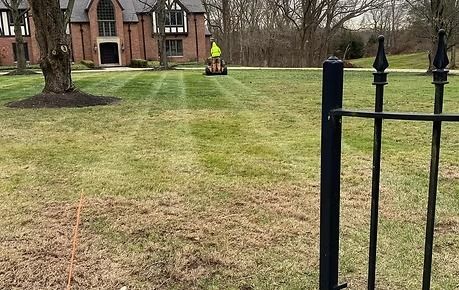 Grassy lawn with a black fence in foreground and a person in a neon vest near houses in the background