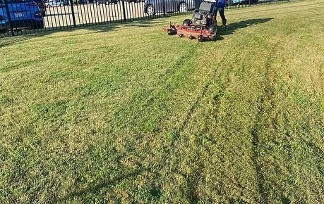 Two workers mow a grassy field with a red riding mower near a fence.