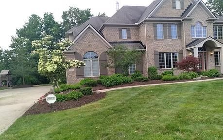 Large brick suburban house with manicured lawn, shrubs, and driveway under cloudy sky