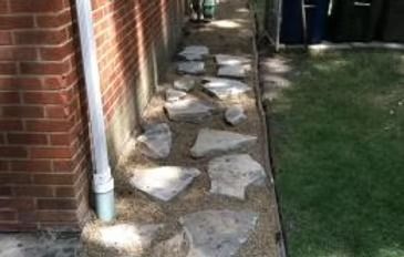 Stone stepping path beside a brick house, leading past a downspout to a grassy yard
