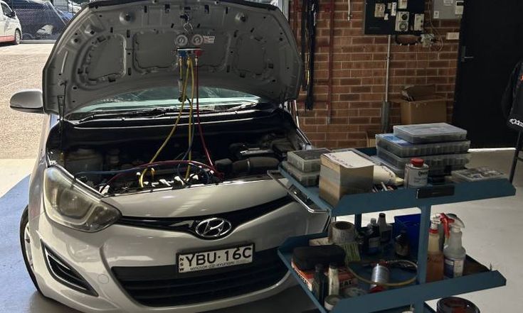 A silver Hyundai car with its hood open in a garage, undergoing air conditioning service.