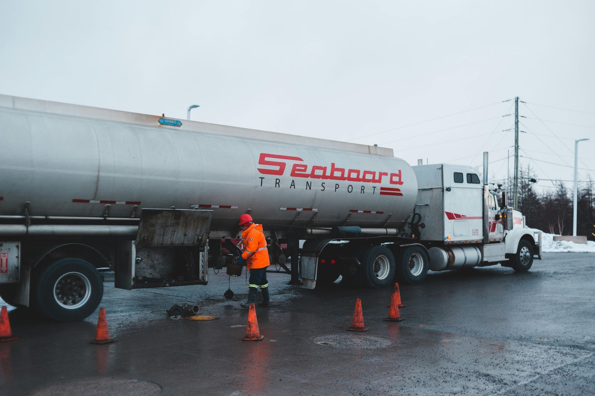 Person in orange safety gear near a Seaboard Transport tanker truck, surrounded by traffic cones, on asphalt.
