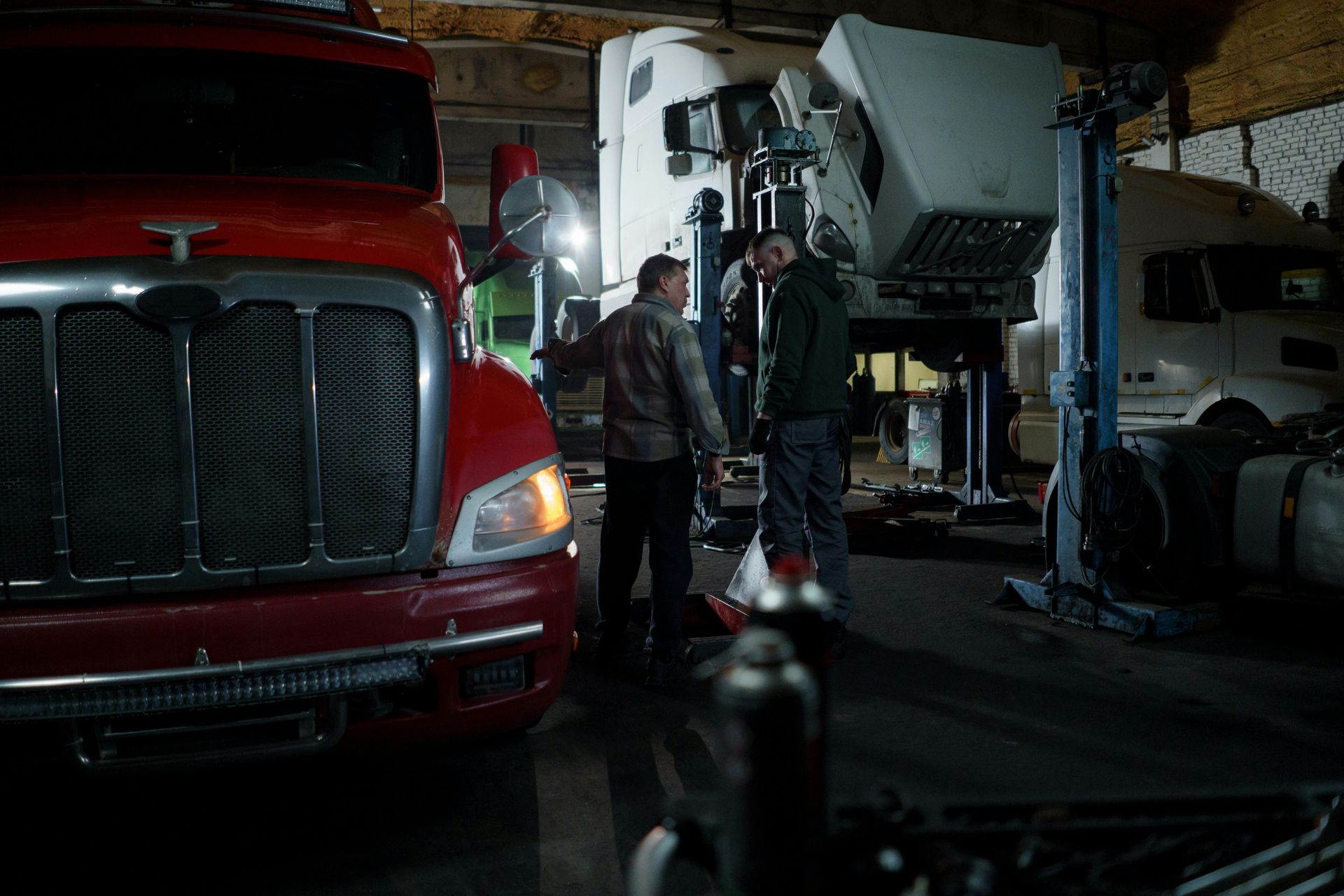 Two men talking in a dimly lit truck repair shop, red truck in foreground, white truck elevated.