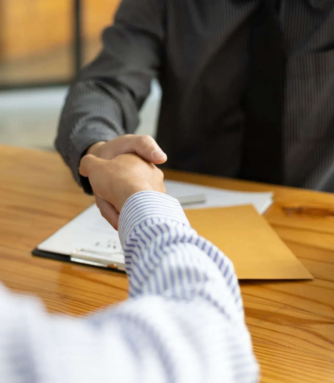 Two men shaking hands over a wooden table, paperwork and a manila envelope present.