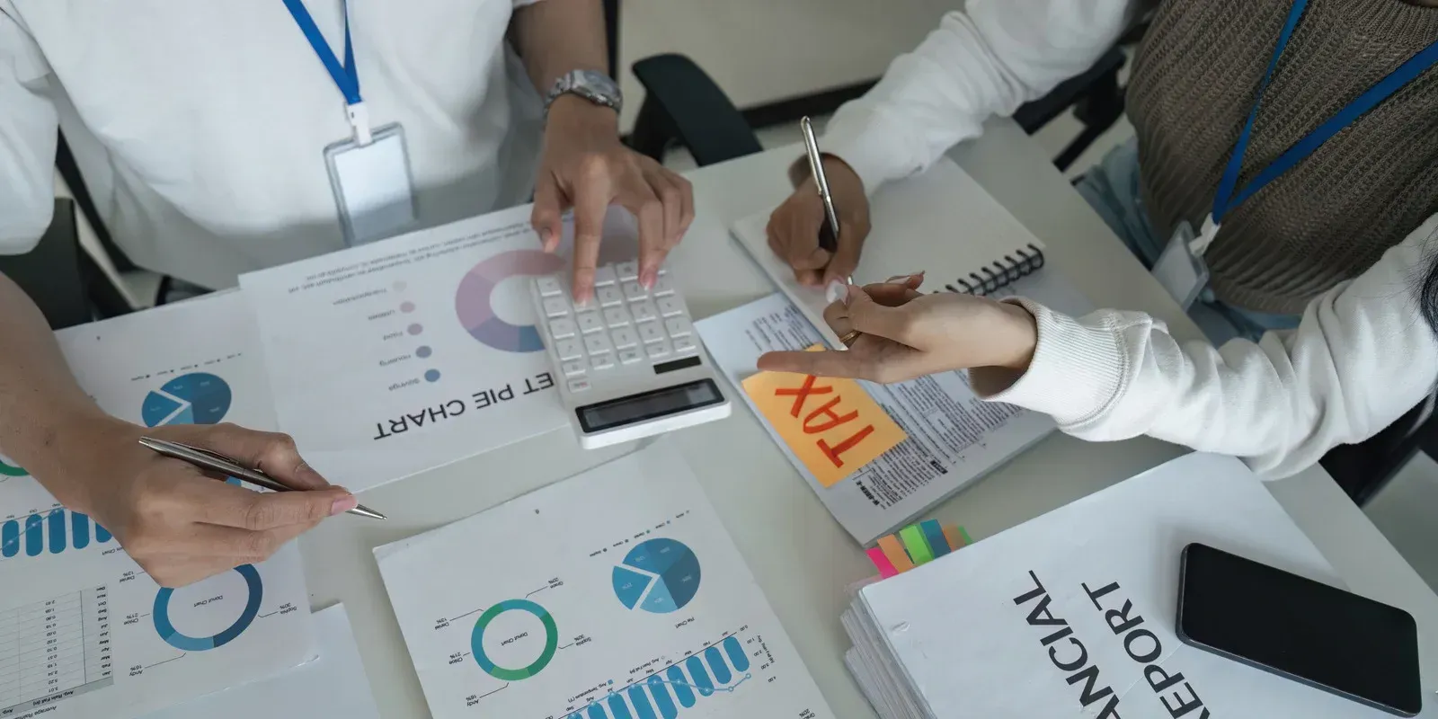 Two people reviewing financial reports at a desk. One uses a calculator, the other writes notes.