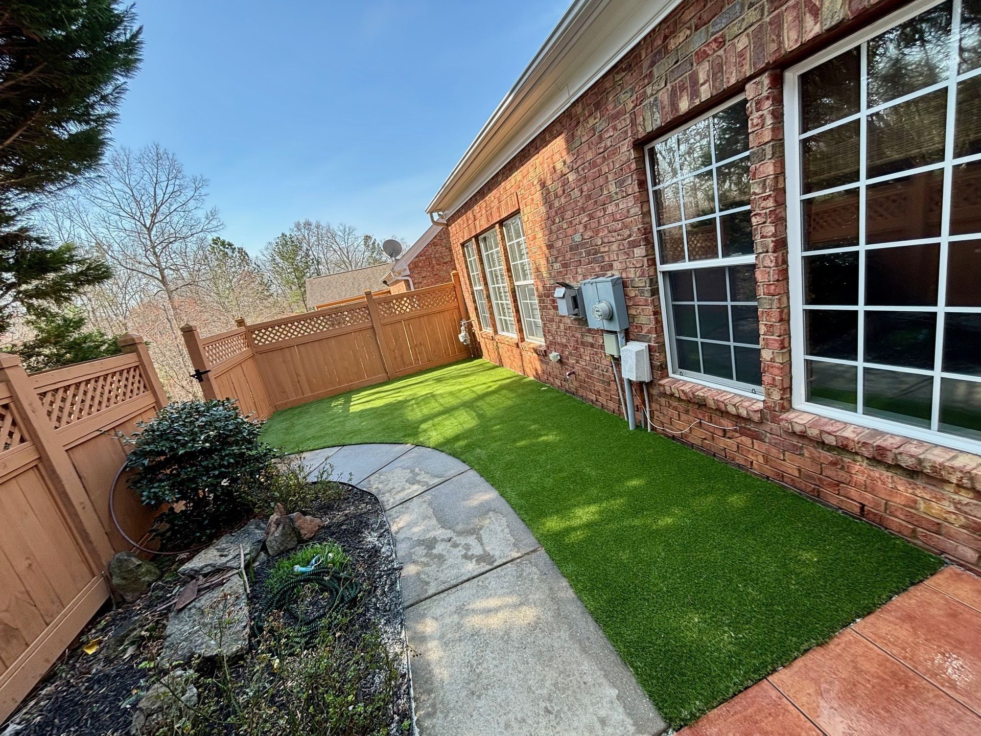 Small backyard with green turf, brick house, stone path, and wooden fence. Sunny day.