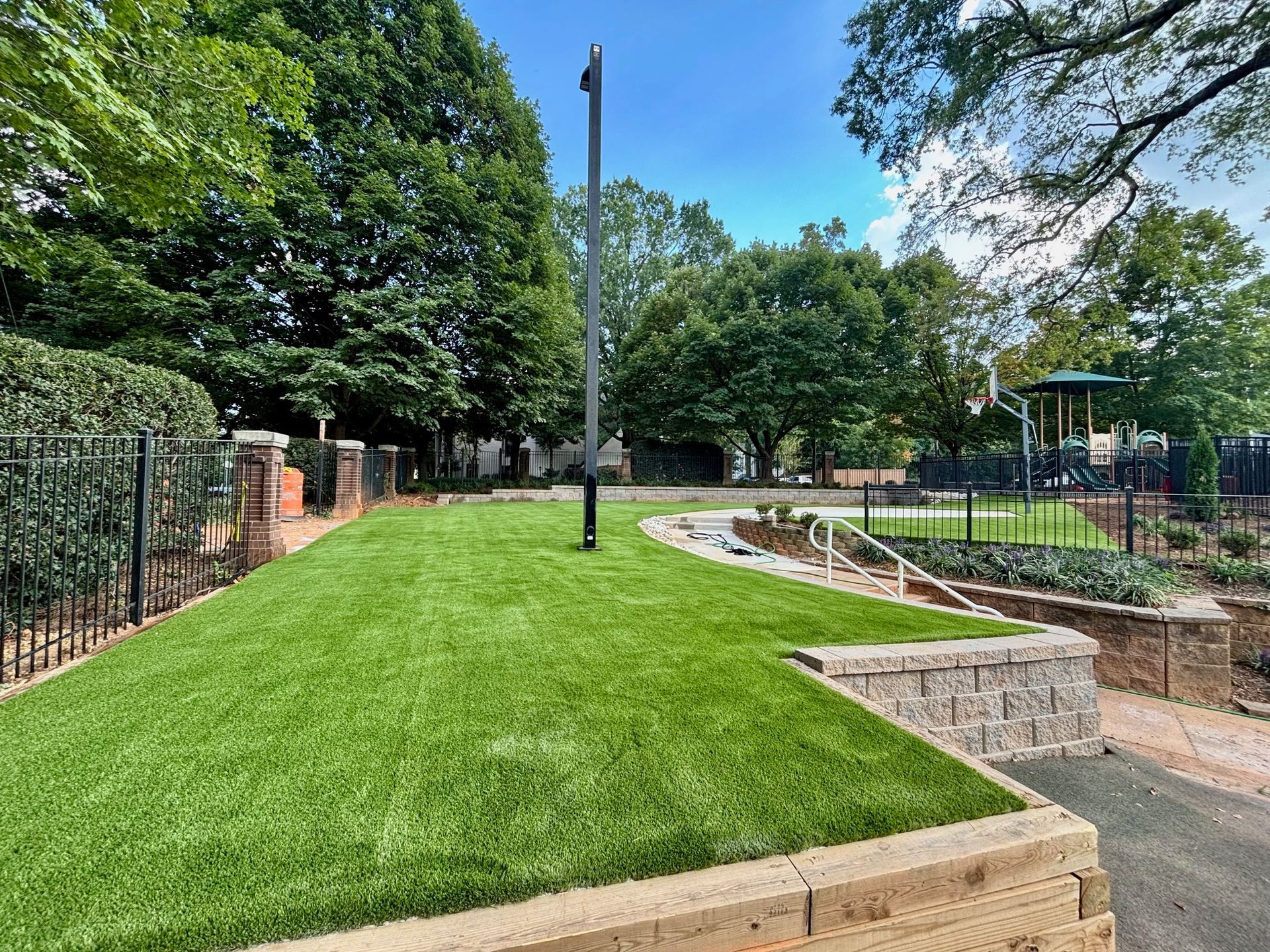 Green artificial turf with a metal pole, brick wall and trees in a park.