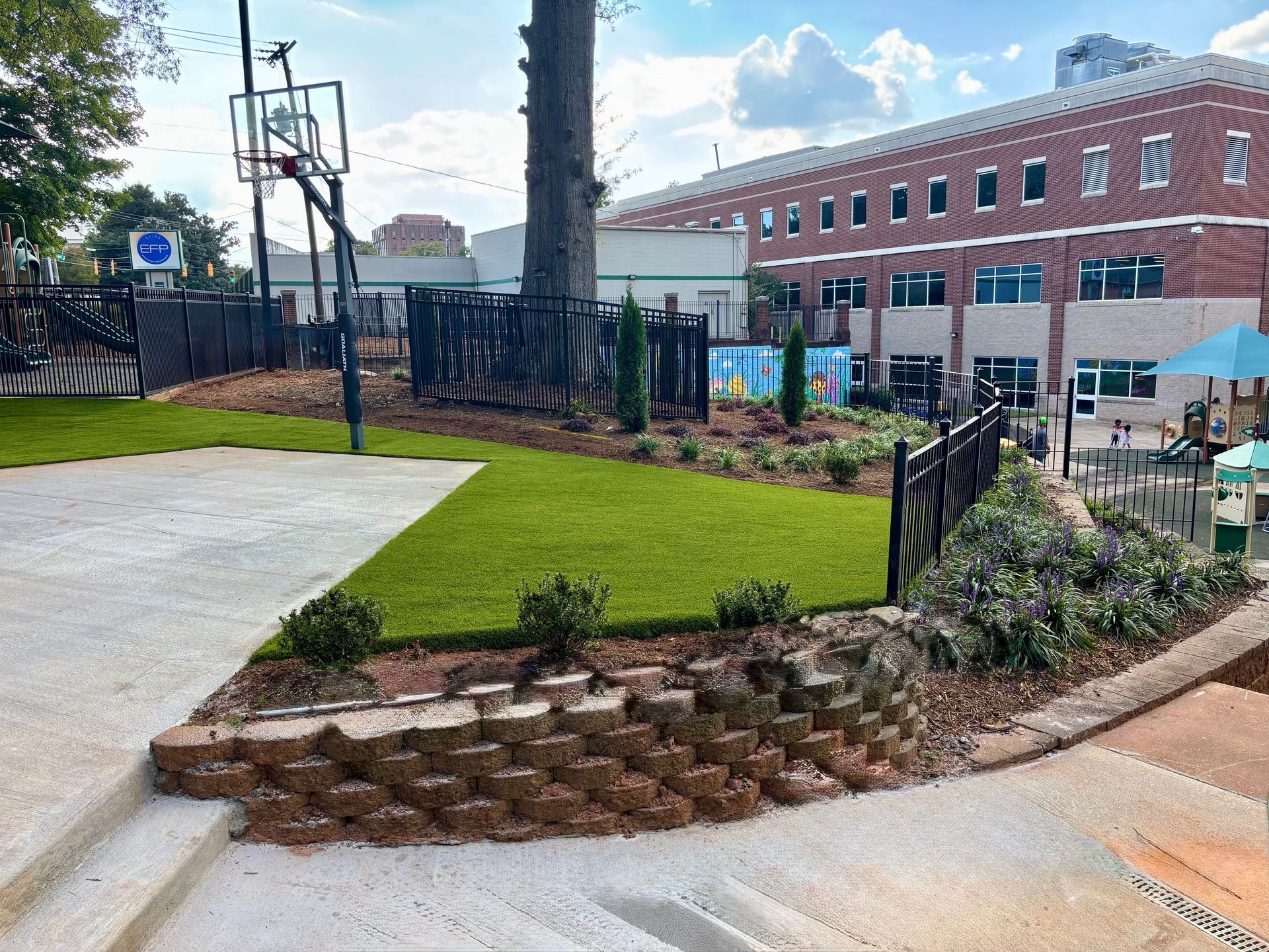 Basketball court with artificial grass, brick retaining wall, and building in the background.