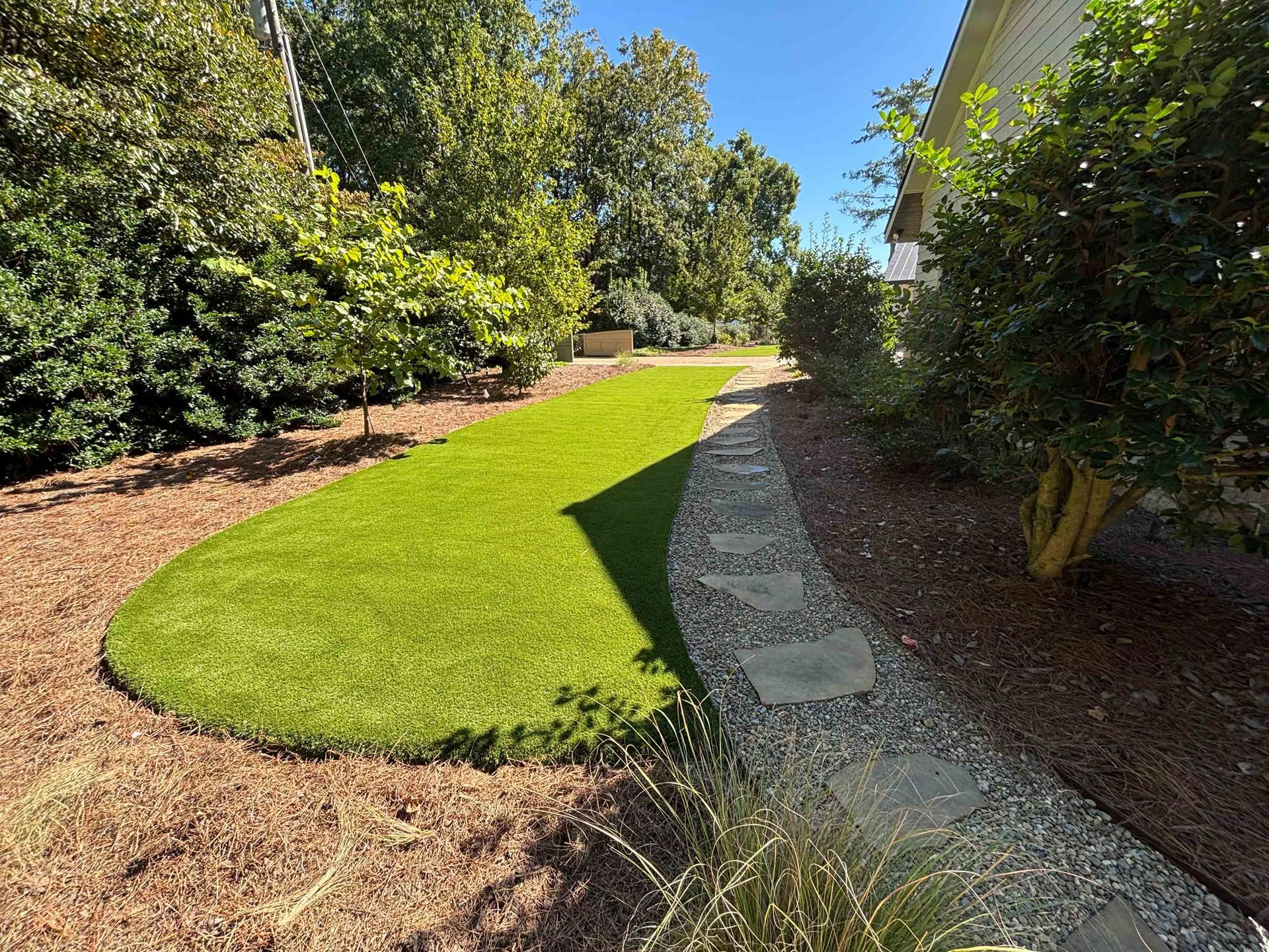 Green artificial lawn beside a stone path, surrounded by trees and bushes. Bright sunny day.