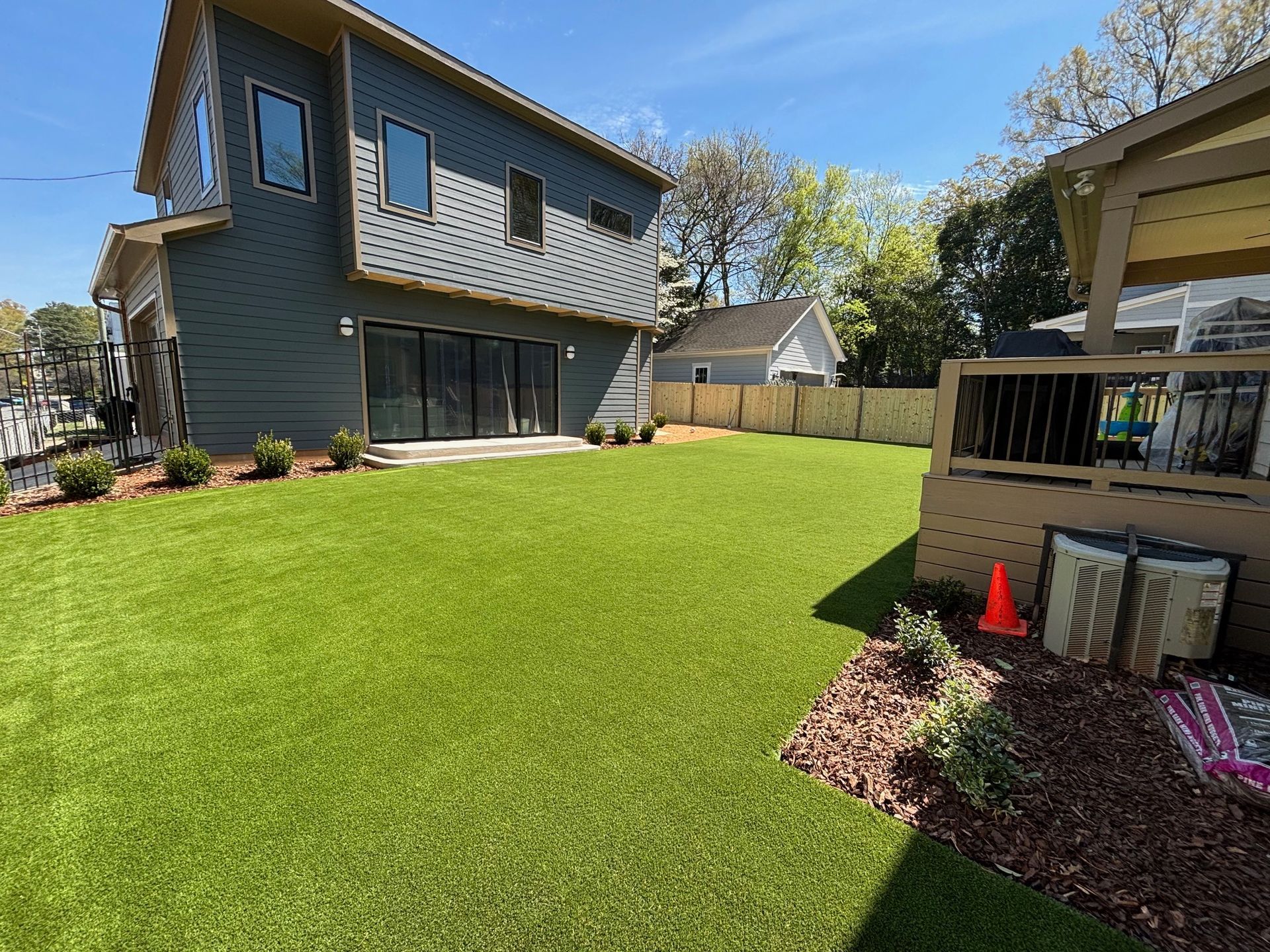 A backyard with vibrant green artificial turf, a gray two-story house, a wooden deck, and a small area of brown mulch.