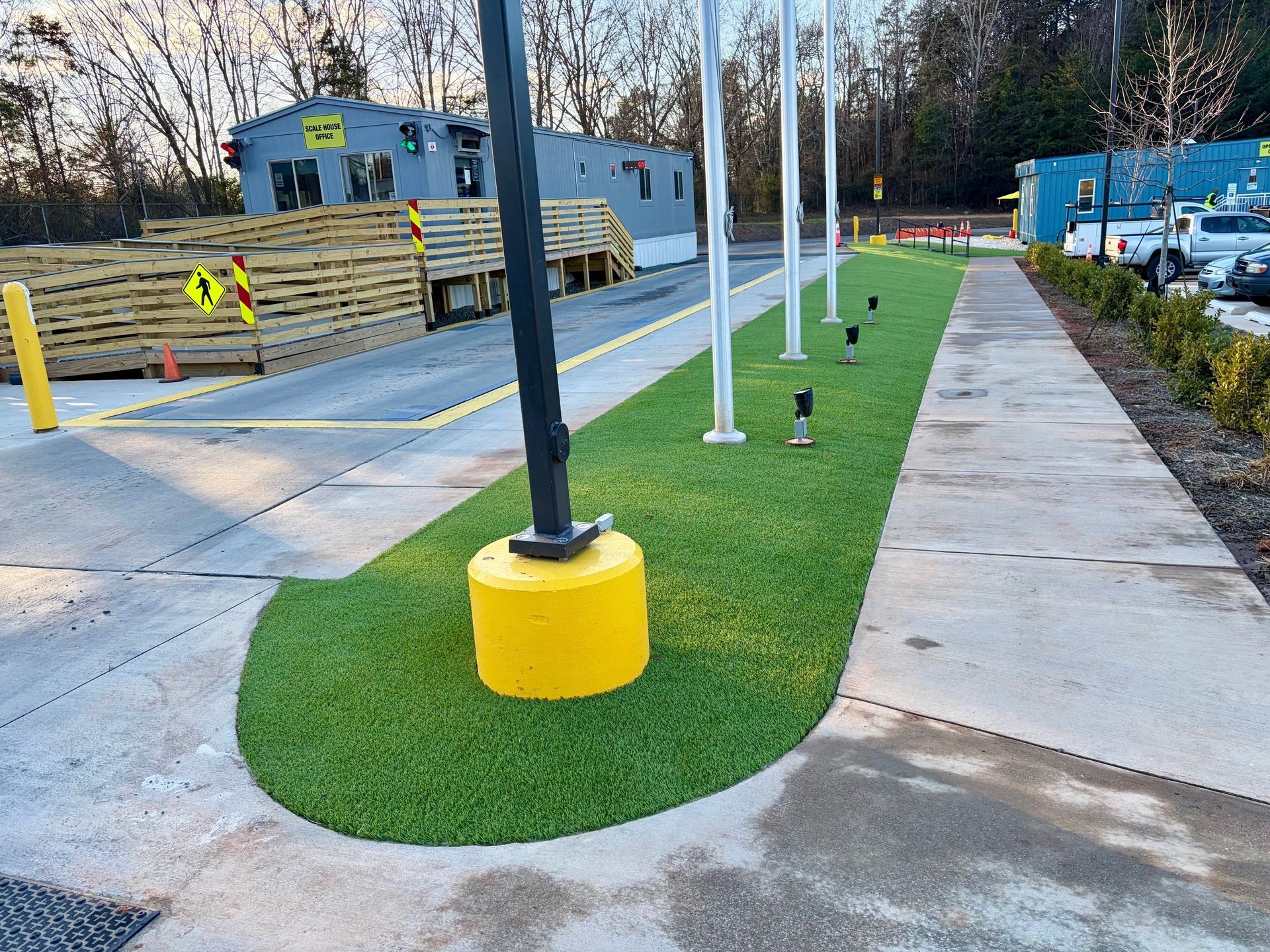A sidewalk with a strip of artificial turf, flag poles, and a yellow base for a pole.