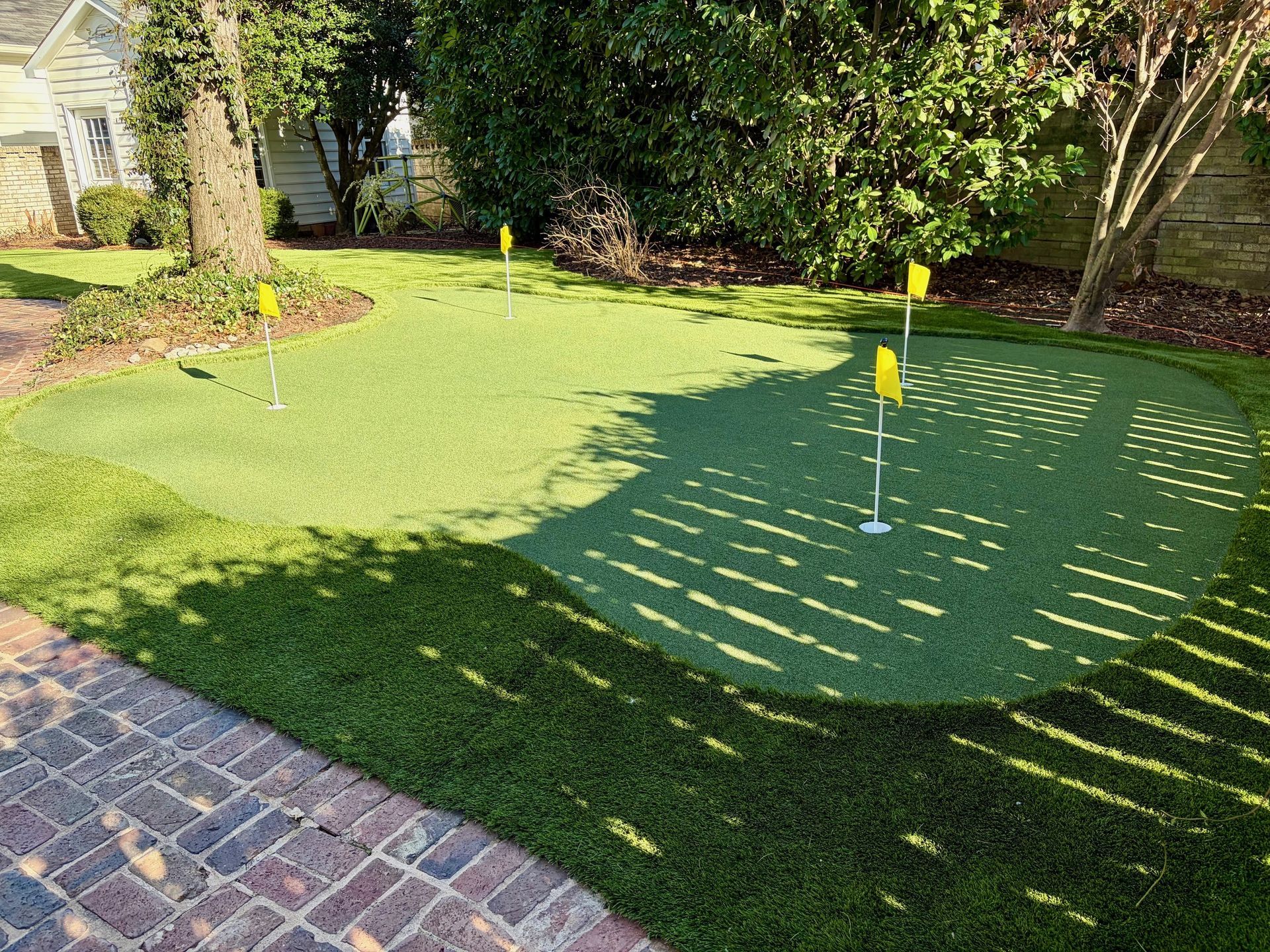 Backyard putting green with three holes and yellow flags, surrounded by grass and trees.