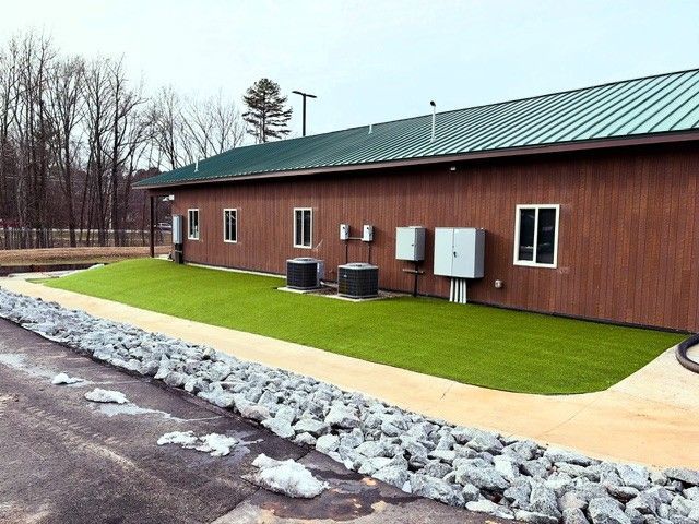 Brown building with green metal roof, artificial turf, and rock border alongside a parking area.