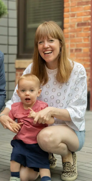 A woman is kneeling down next to a baby and smiling.