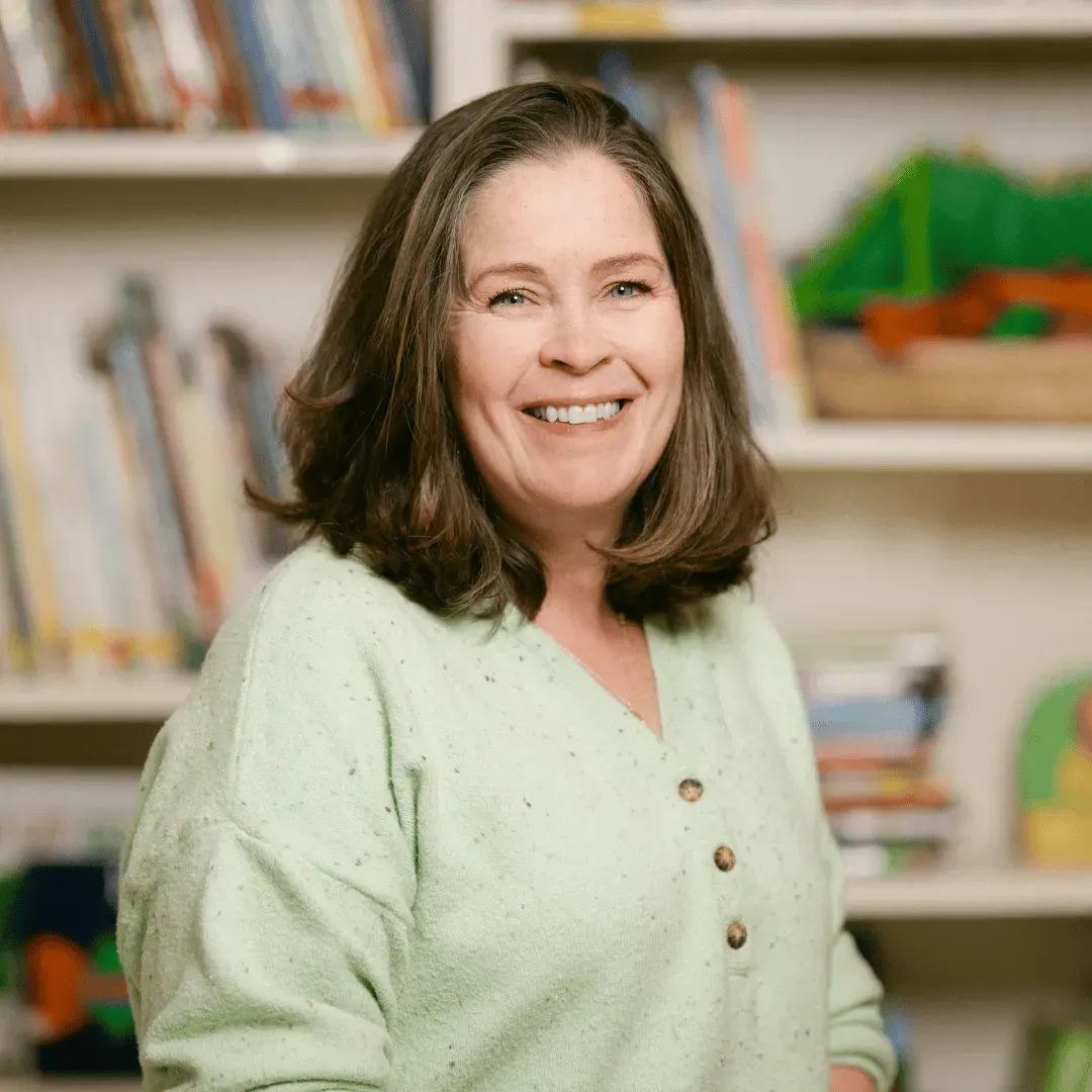 A woman in a green sweater is smiling in front of a bookshelf.