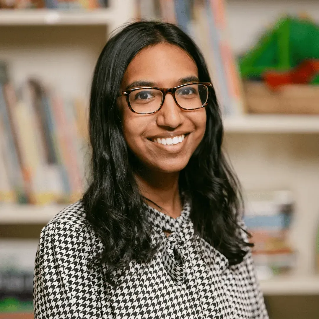 A woman wearing glasses and a plaid shirt is smiling in front of a bookshelf.
