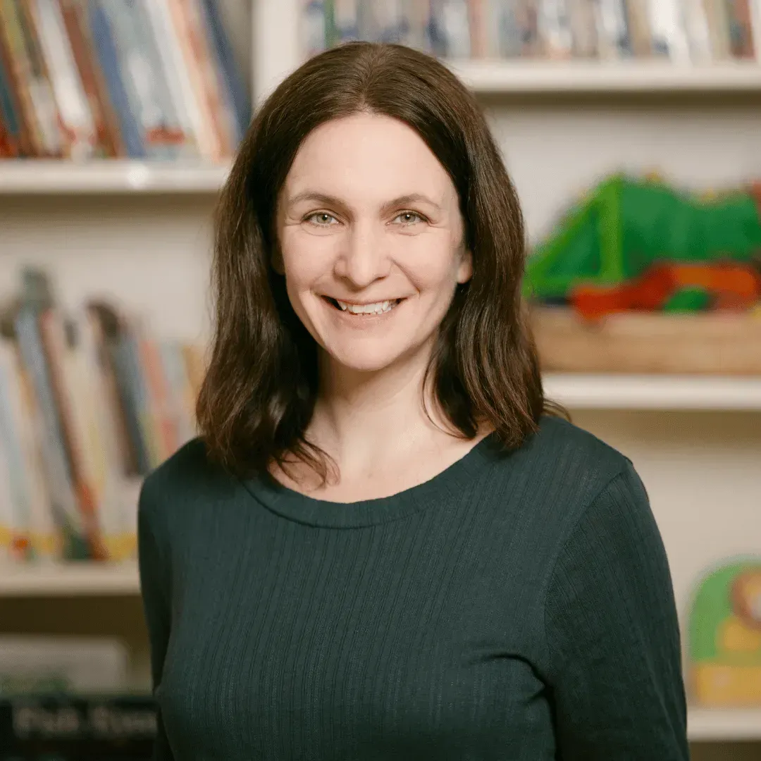 A woman in a green shirt is smiling in front of a bookshelf.
