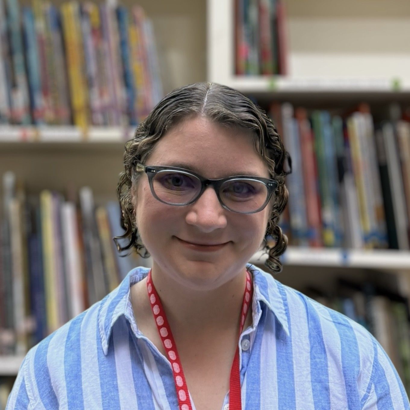 A woman wearing glasses and a lanyard is smiling in front of a bookshelf.