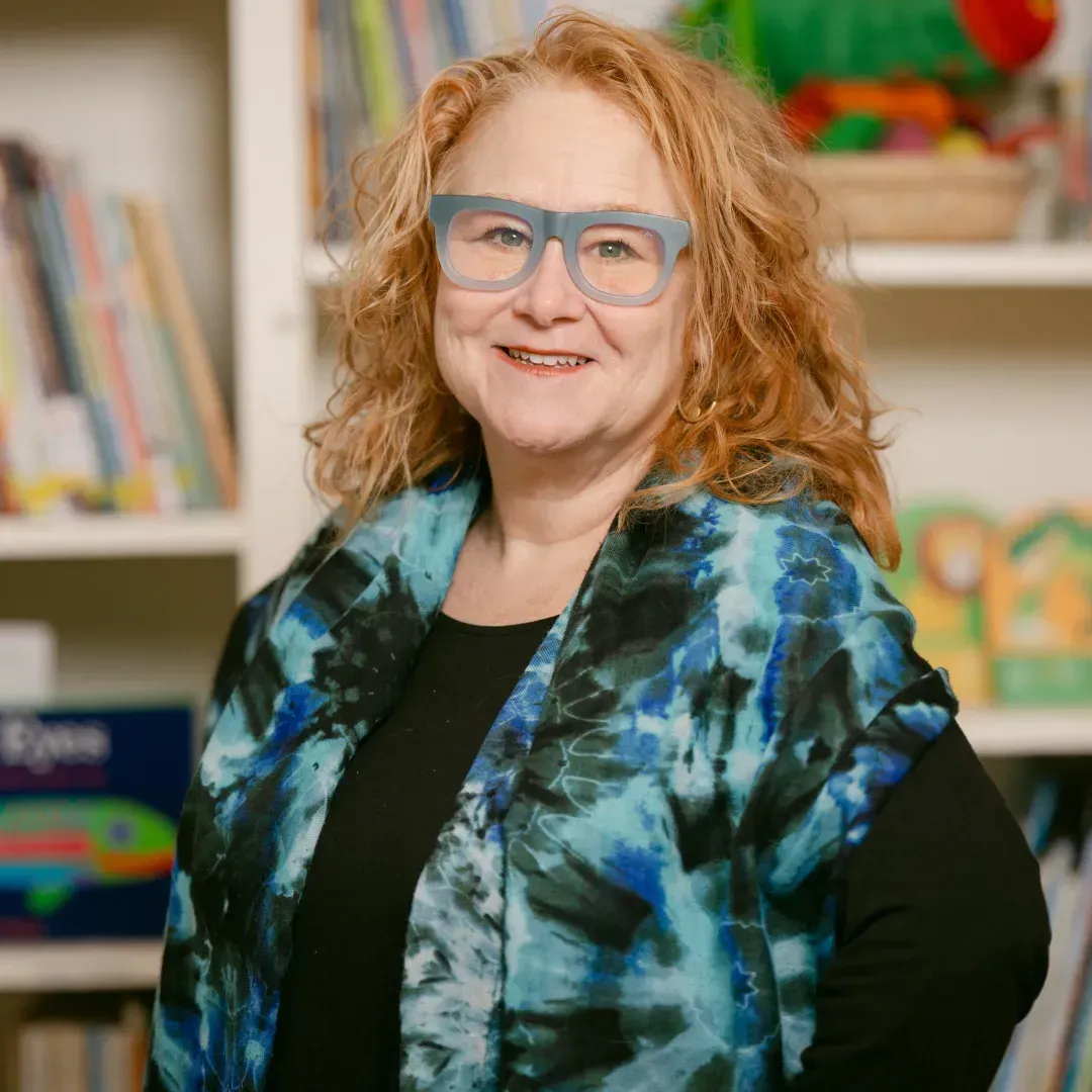 A woman wearing glasses and a blue scarf is standing in front of a bookshelf.