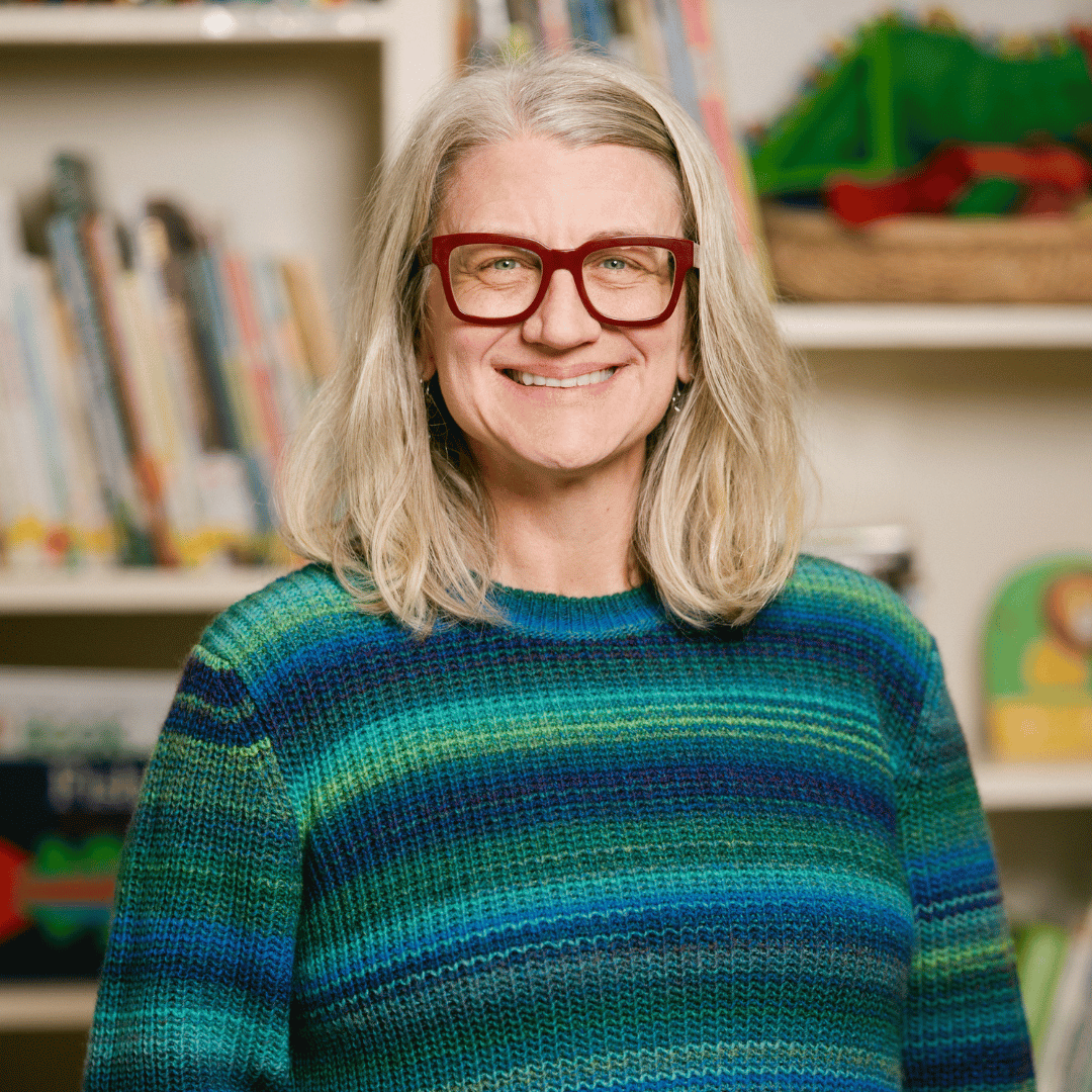 A woman wearing glasses and a blue and green sweater is smiling in front of a bookshelf.