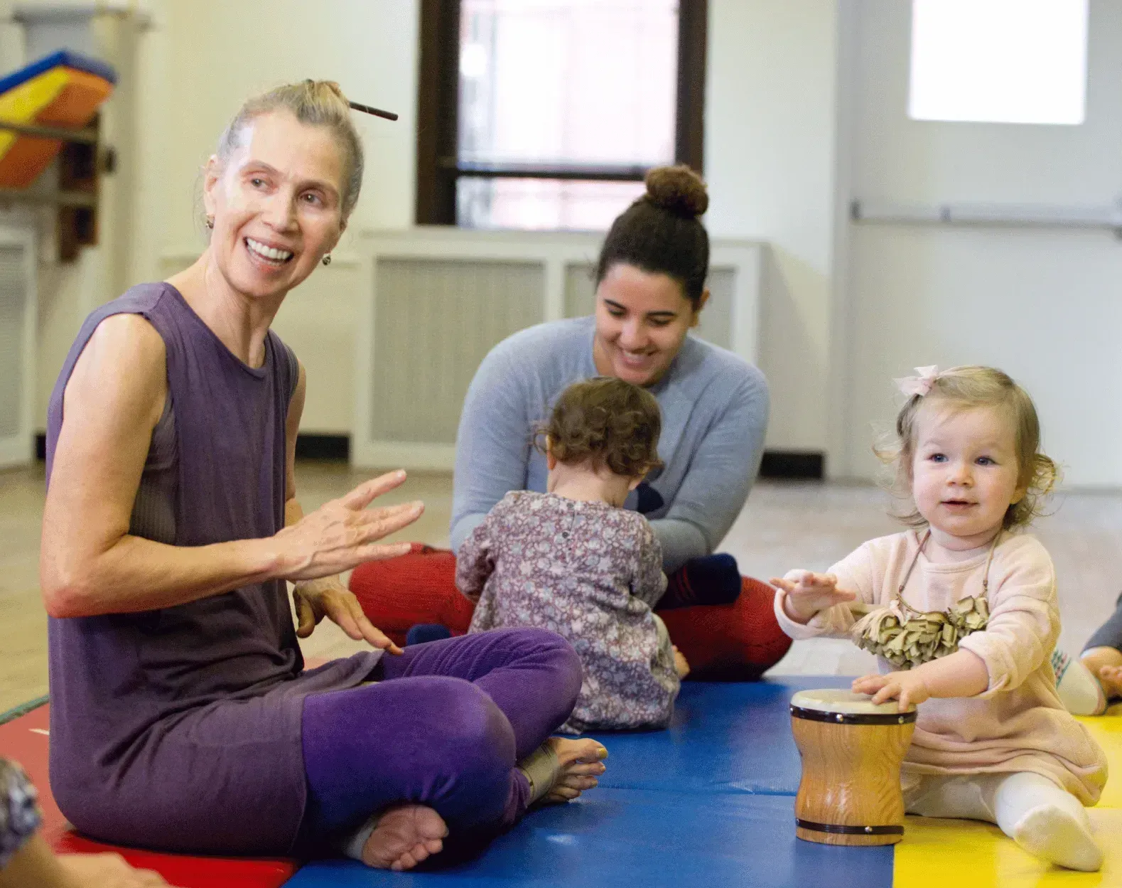 A woman is sitting on the floor with two babies and a drum.