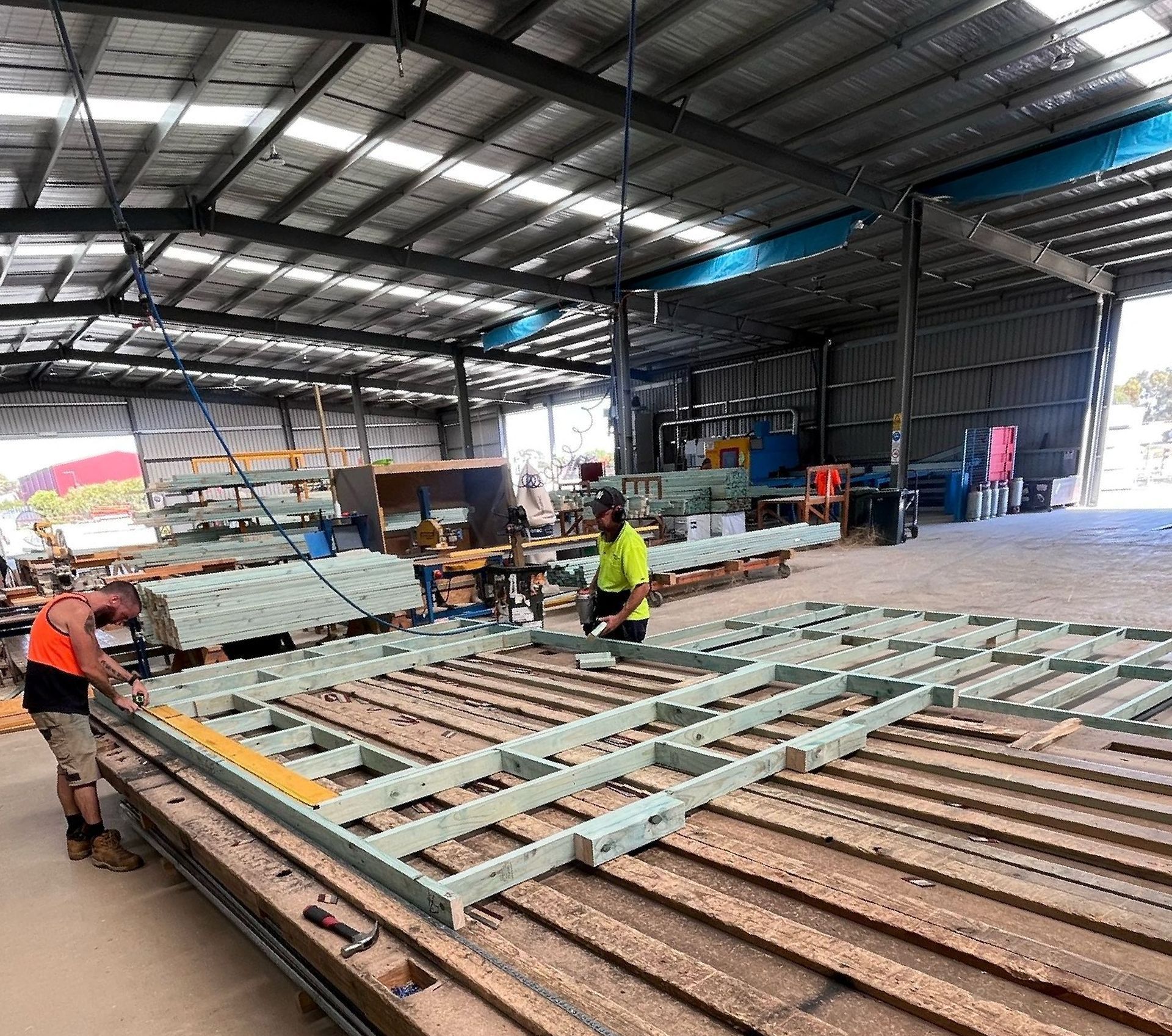 Two construction workers assembling a metal frame inside a large warehouse with building materials scattered around.