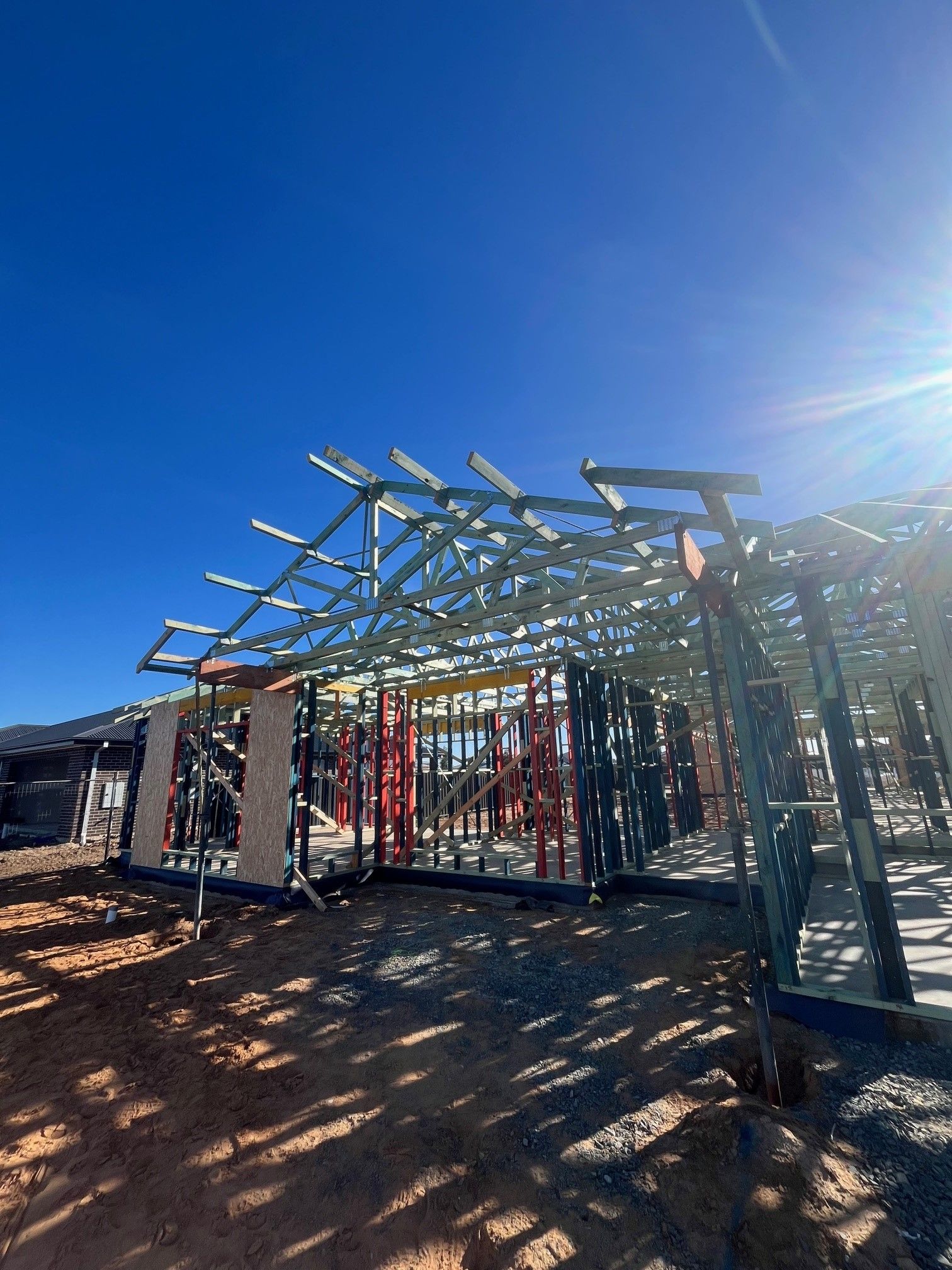 Steel frame of a house under construction against a clear blue sky. The sun shines brightly, and the ground is dirt.