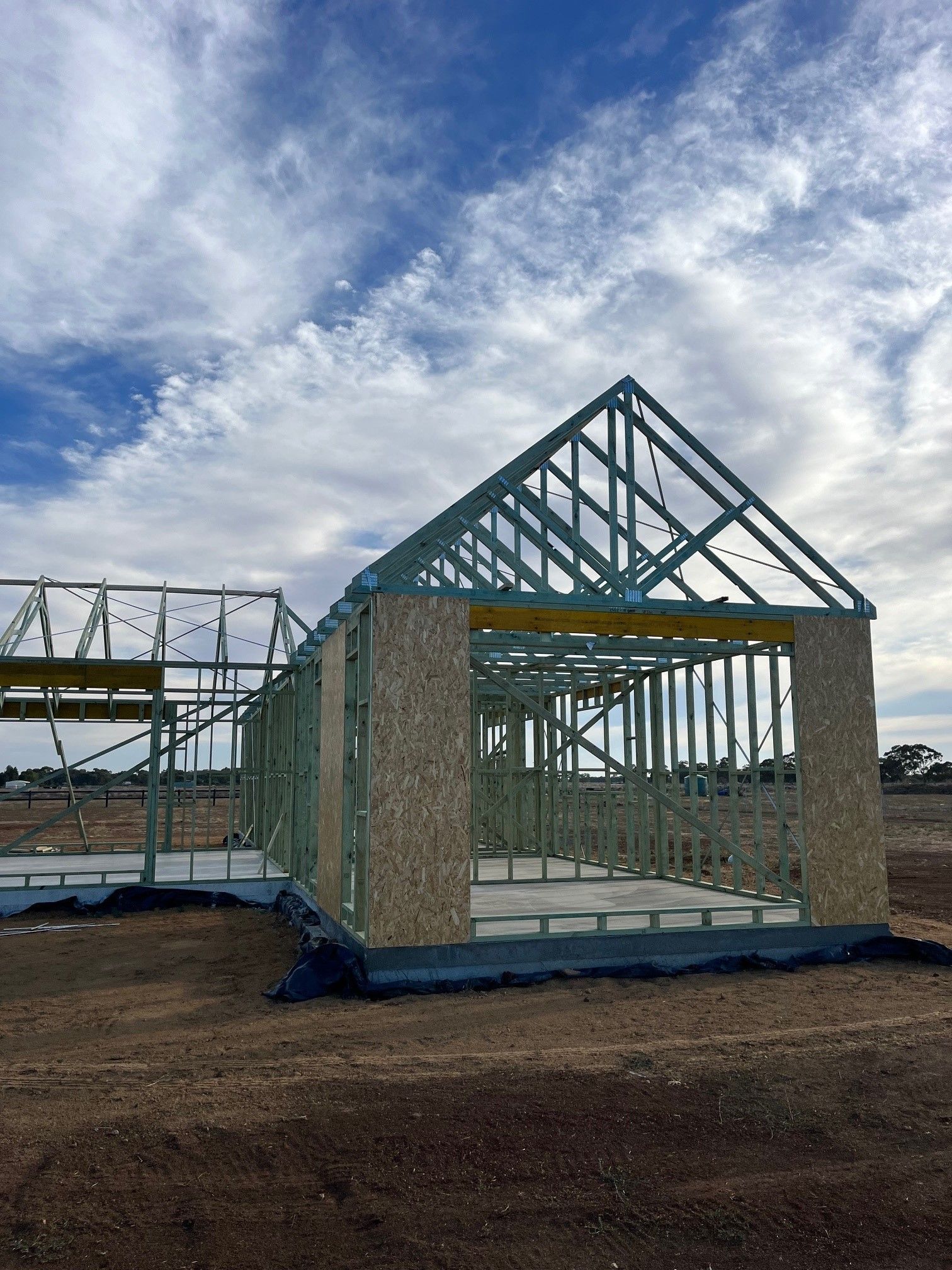 Framed structure of a house under construction against a cloudy blue sky, on a dirt lot. 