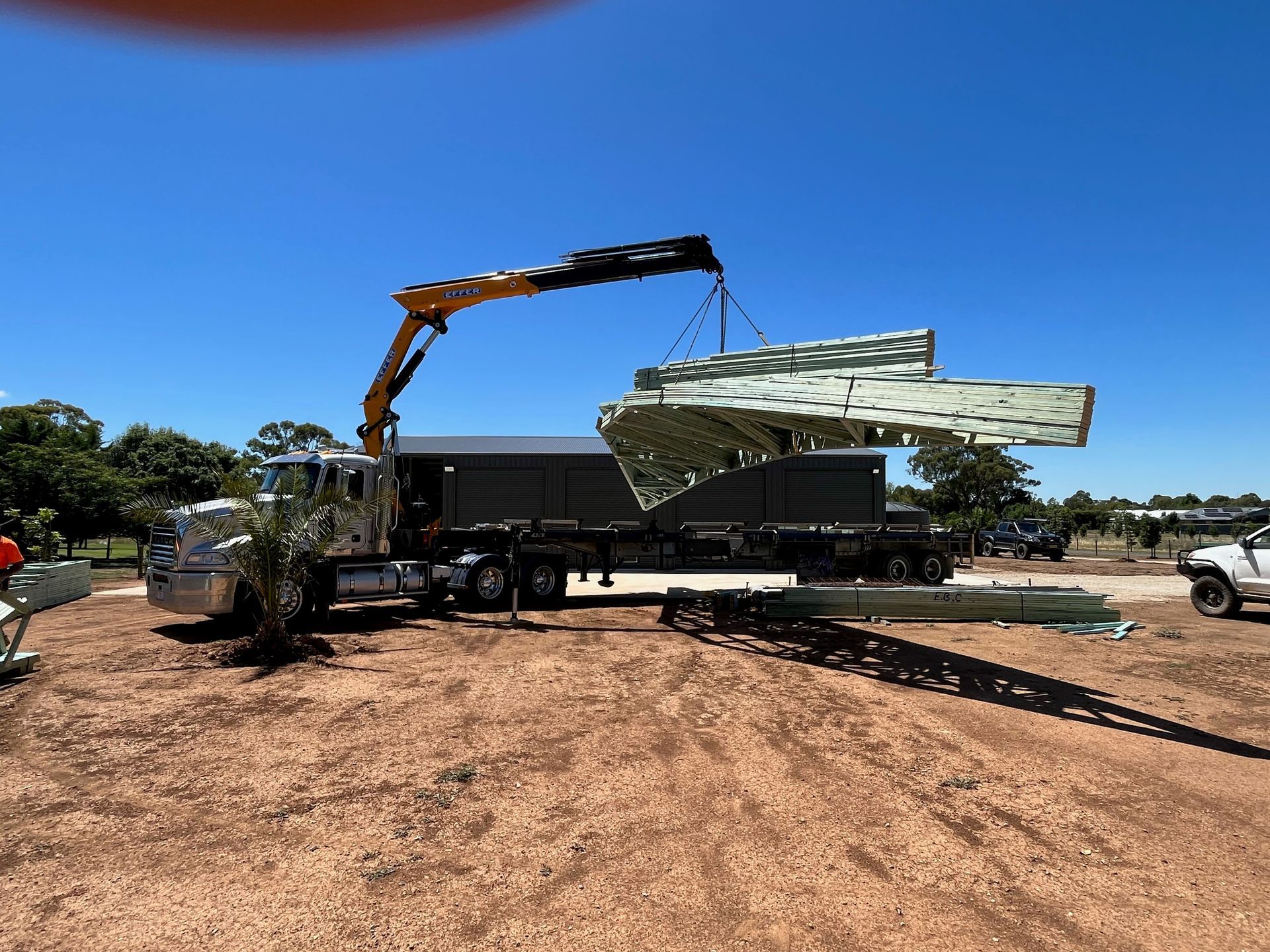 A crane truck lifting large panels from a flatbed trailer on a construction site under a blue sky.