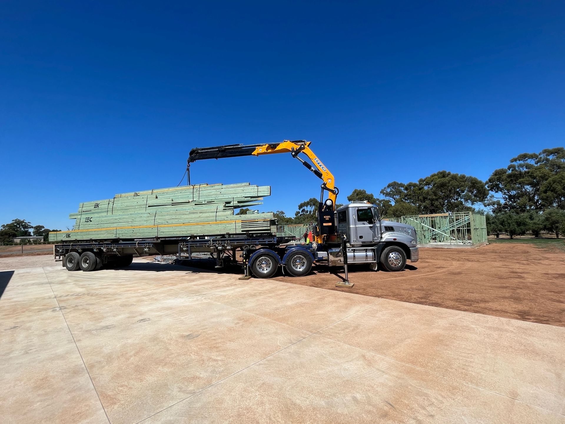 A crane truck unloading lumber on a sunny day. The truck is silver and the sky is bright blue.