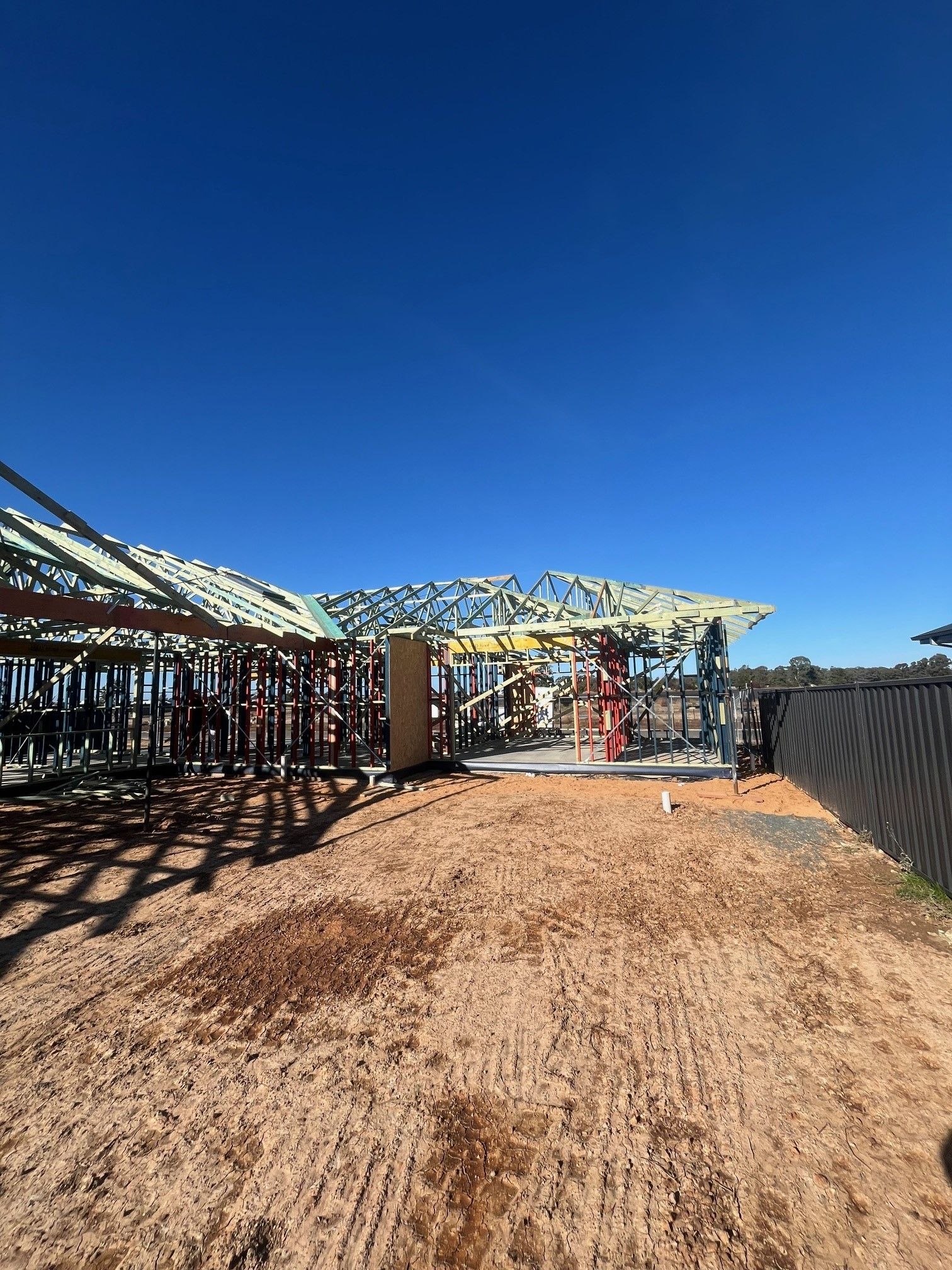 Construction site of a house under a clear blue sky. The framing of the roof and walls are visible on a dirt lot.