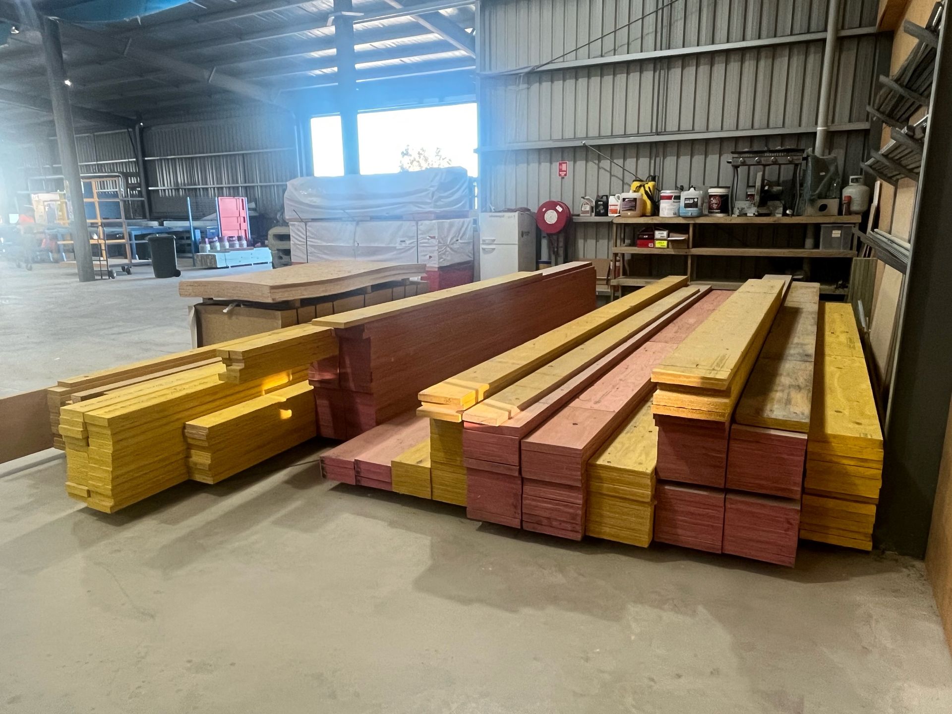 Warehouse interior with stacks of lumber in yellow and reddish-brown hues, near a window and shelves with tools.