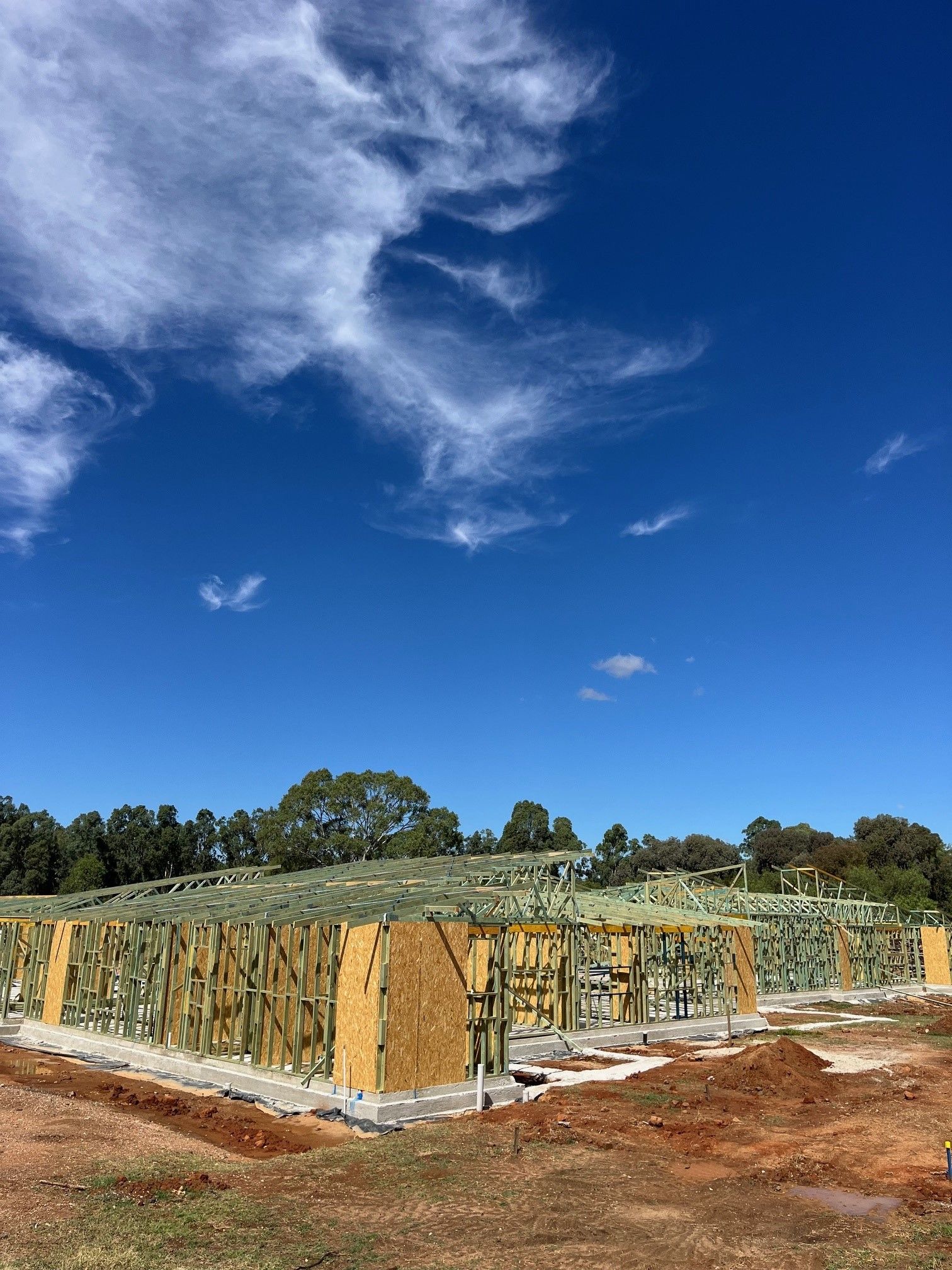 Framing of a building under construction against a bright blue sky with wispy clouds; dirt ground in foreground.