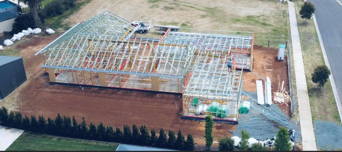 Aerial view of a house under construction. Wooden framing is visible, set on a dirt lot next to a road.