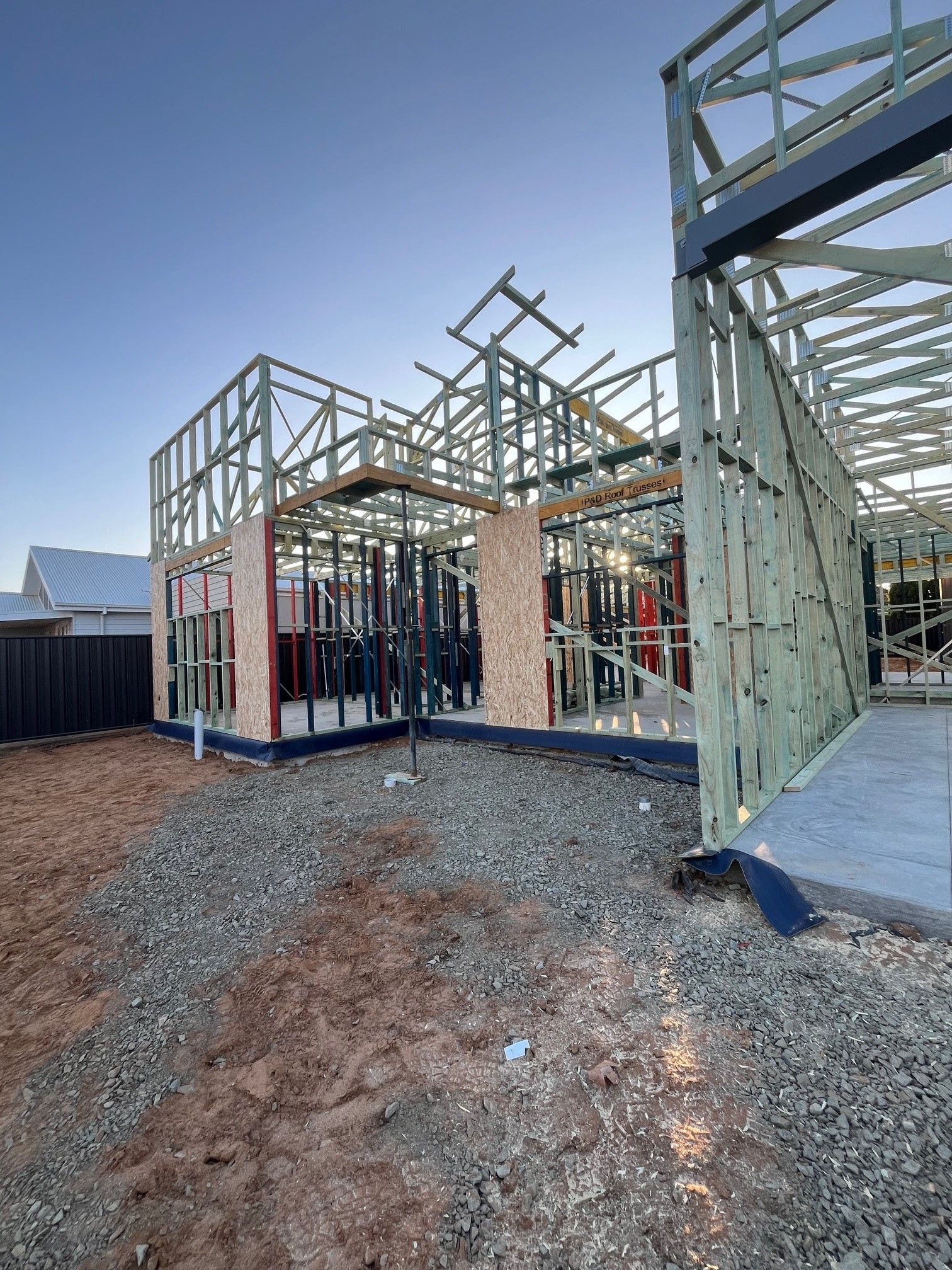 A partially constructed house frame with wooden beams, set on a gravel area, under a clear blue sky.