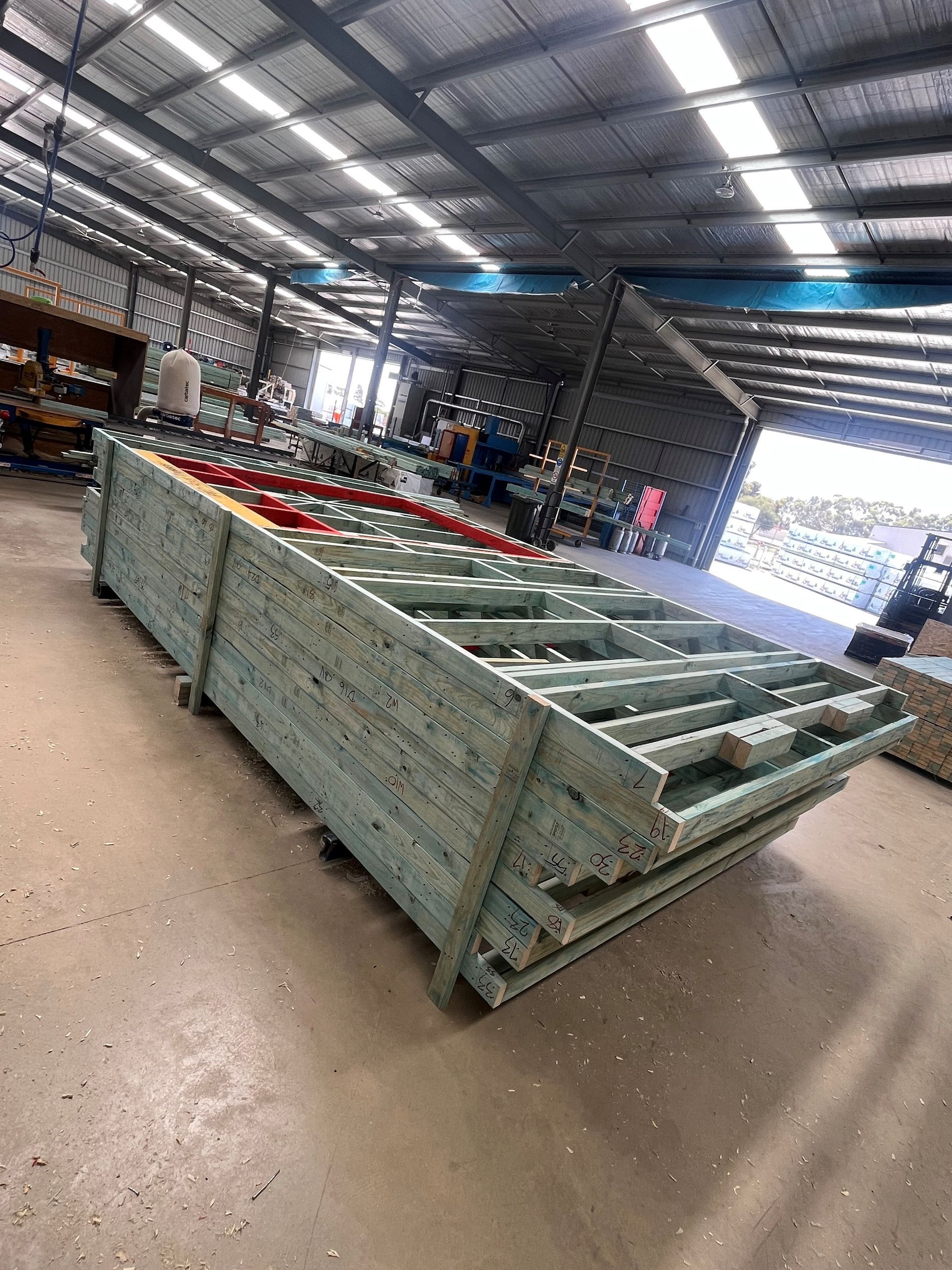 A stack of wooden wall frames in an industrial warehouse. The frames are green, and the ceiling has metal beams.