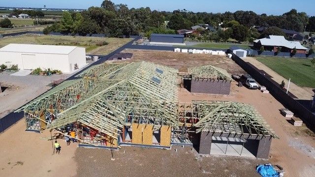 Aerial view of a house under construction; wooden frame with exposed roof trusses, workers on site, suburban setting.