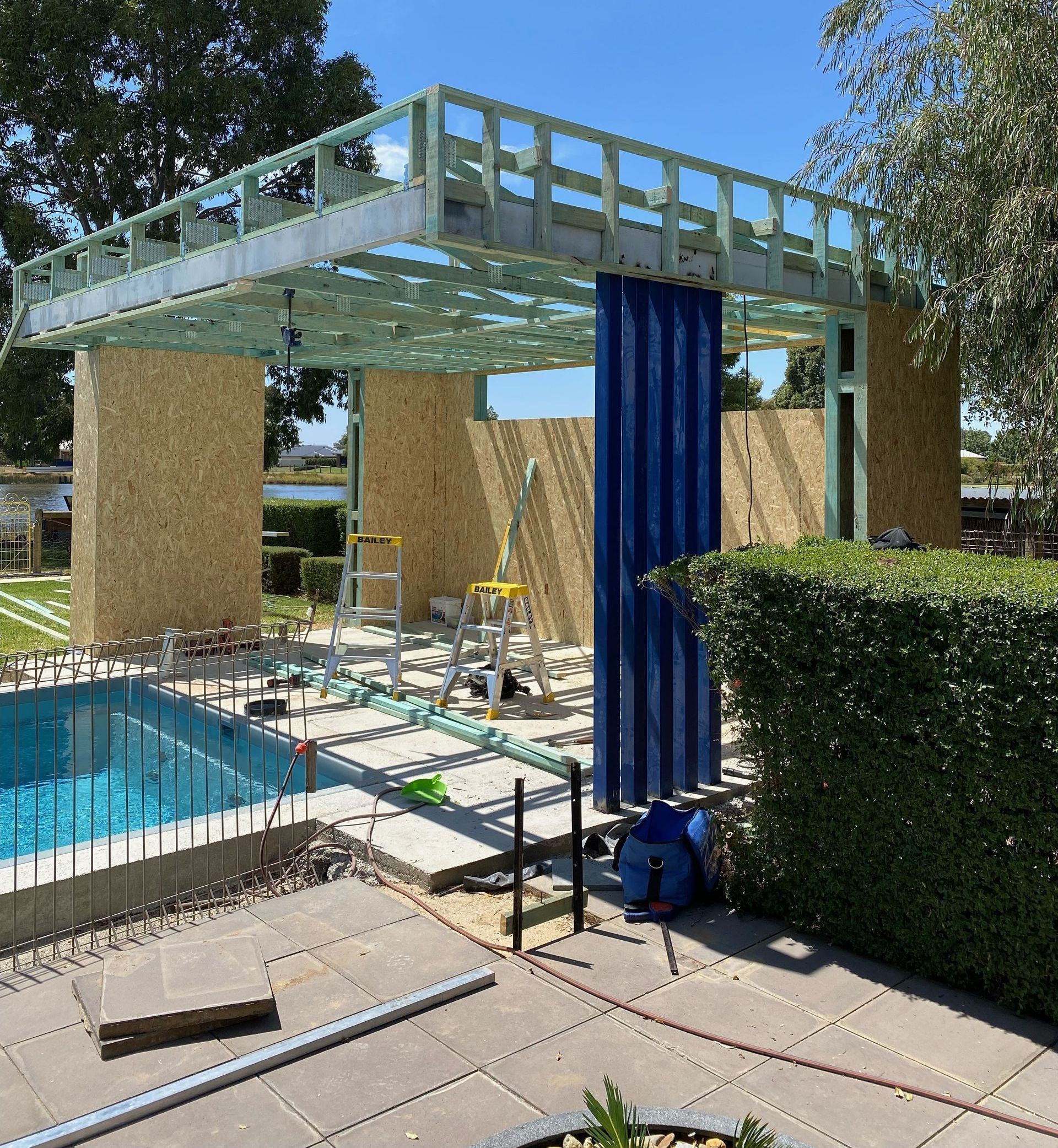 Construction of a poolside cabana.  The frame is up, walls are partially sheathed with wood, and a blue curtain hangs.