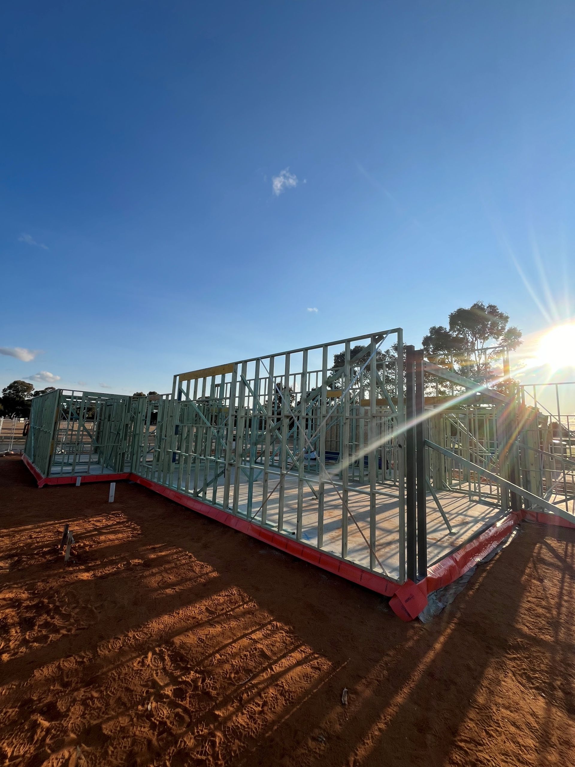 Wooden frame of a house under construction against a bright blue sky with the sun shining. Red foundation border.